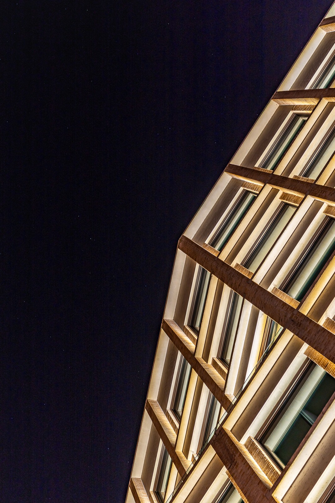 A contemporary facade cut diagonally against the night — warm, illuminated geometry on one side, deep black on the other. Structure reduced to rhythm, light, and the edge between them.