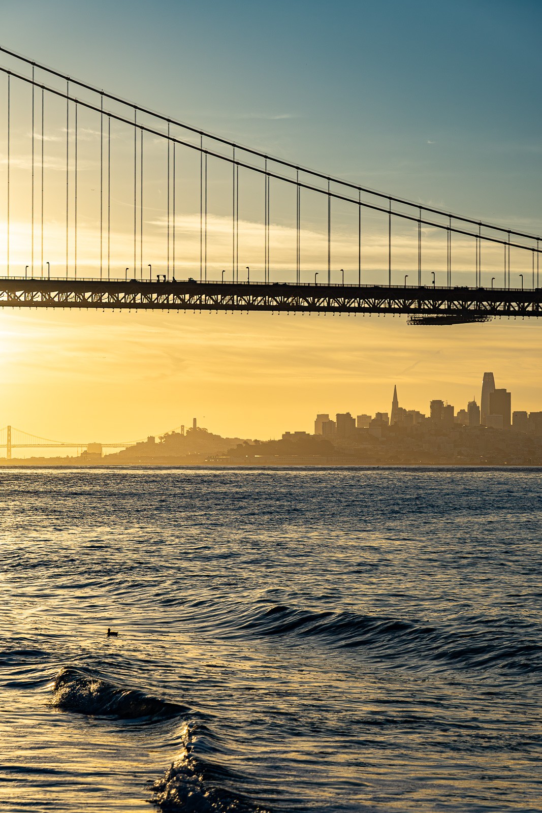 The Golden Gate Bridge seen from Kirby Cove at dusk, San Francisco's skyline silhouetted across the bay. The water is calm, the atmosphere hazy - the bridge and city held at exactly the right distance.