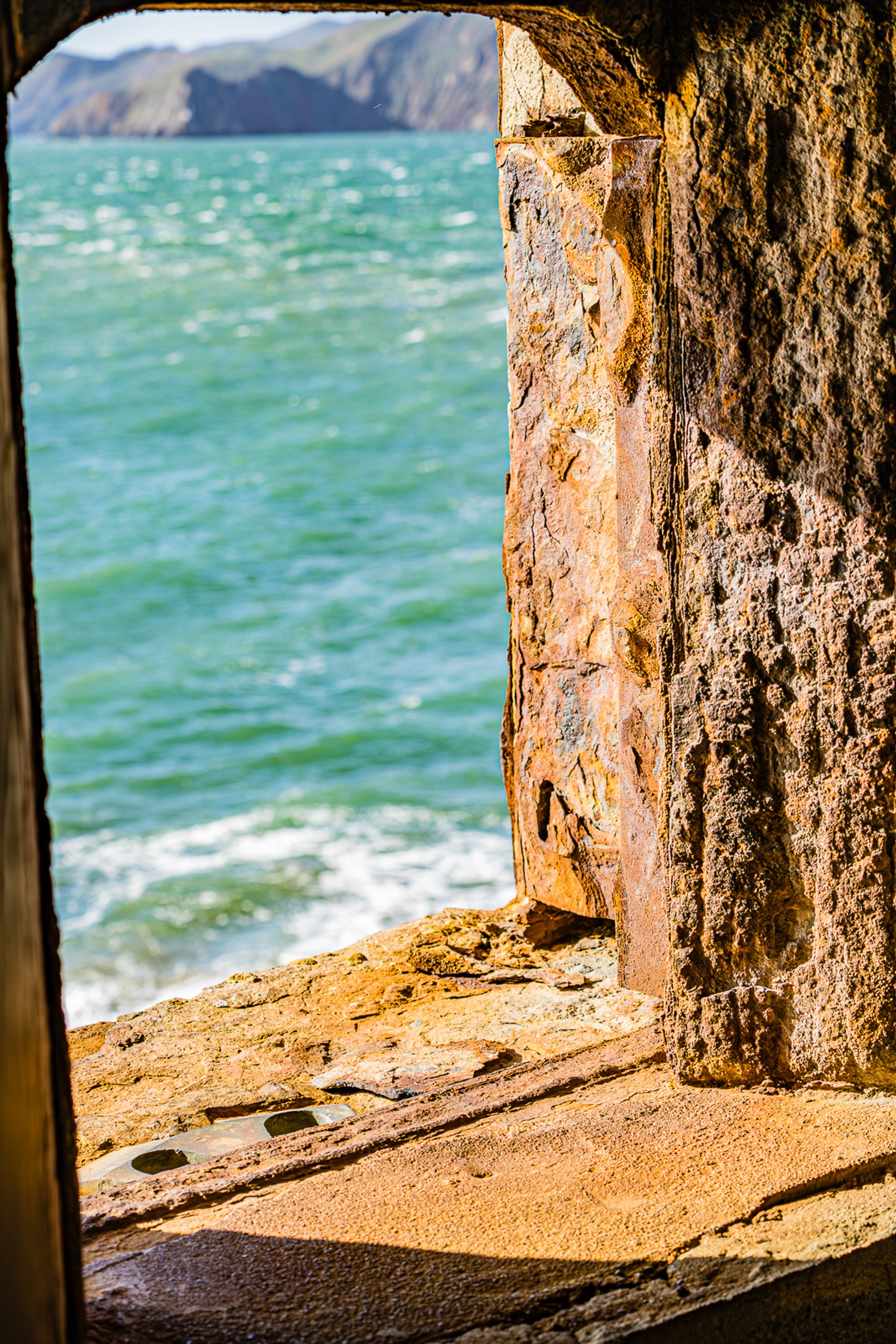 A heavily corroded iron window frame fills both sides of the frame in deep orange and brown rust, and through the opening the Marin Headlands and brilliant turquoise-green bay water are visible below.