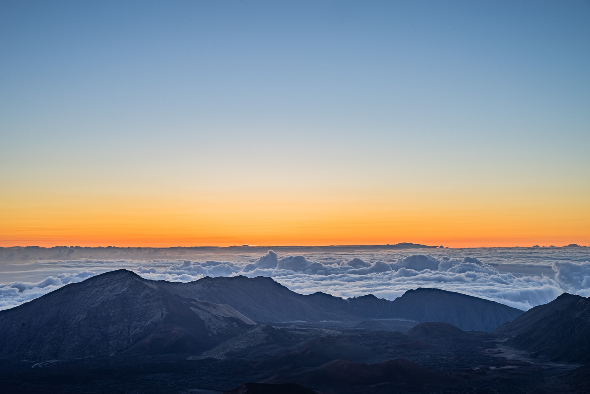 Sunrise from the Haleakalā summit above a complete sea of cloud, distant volcanic peaks emerging through the white layer, the horizon burning orange. An image that requires the 3am alarm and the drive in darkness to reach.
