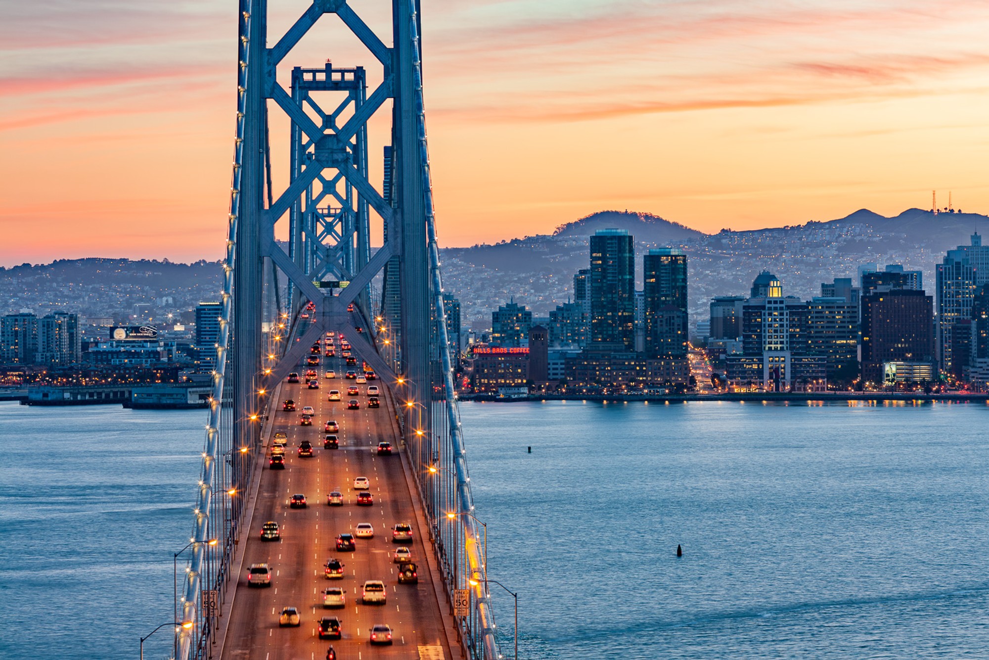 The Bay Bridge at sunset from Yerba Buena Island, the western span silhouetted against a sky of orange and deep pink. The bridge becomes a drawing at this hour - line and colour, with the city beyond.