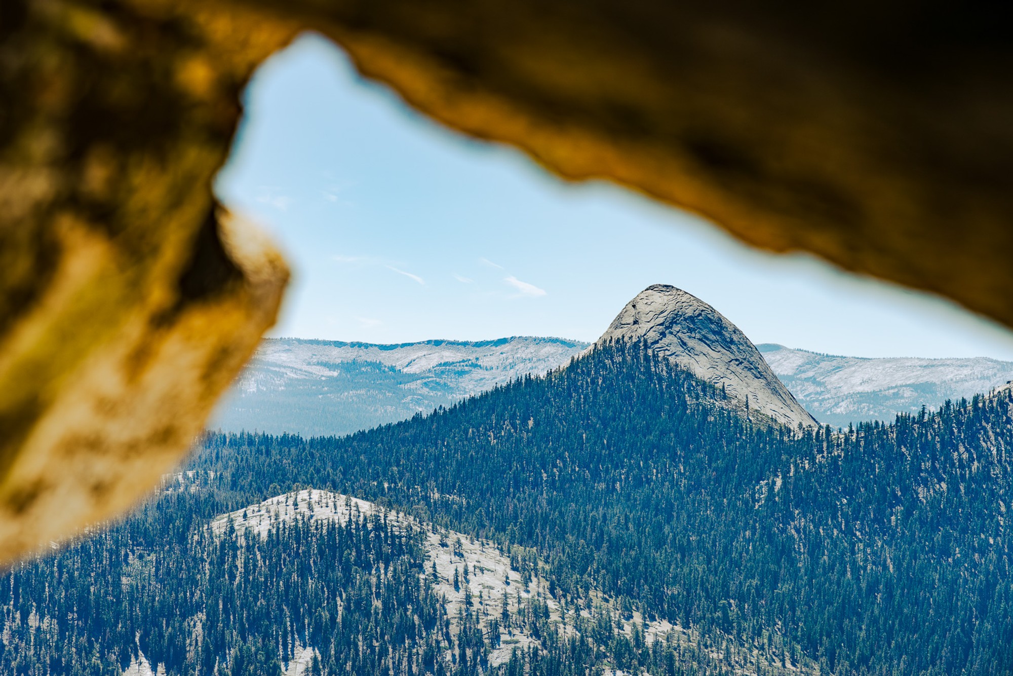 The Yosemite backcountry seen through a natural rock arch, just below Half Dome, the surrounding stone framing the view like a window. The valley floor and forested slopes below