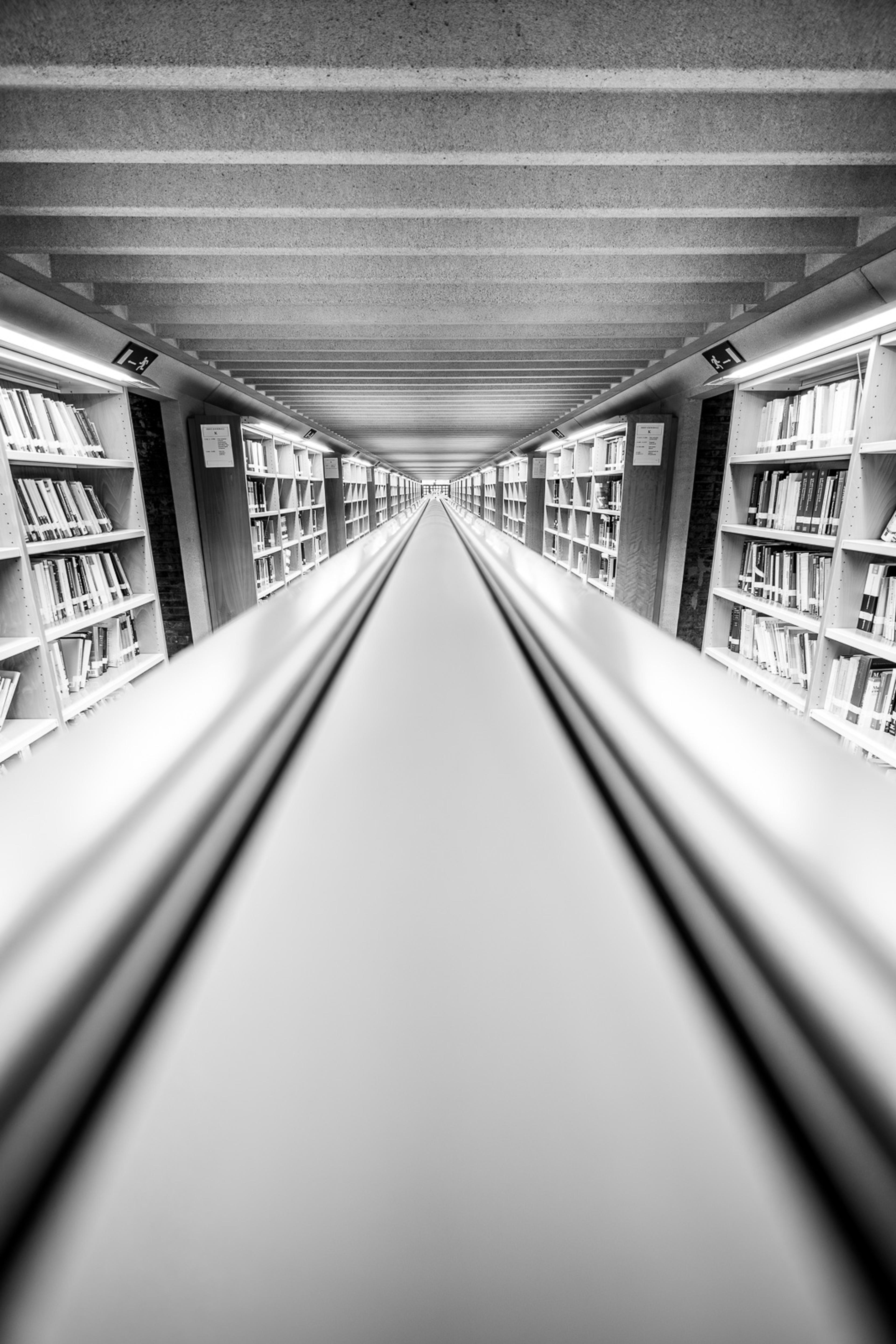 A library corridor in Barcelona shot from one end, right above futuristic reading lights, bookshelves lining both walls to the ceiling as the perspective drives deep toward a white vanishing point. Knowledge as architecture - and the architecture is considerable.