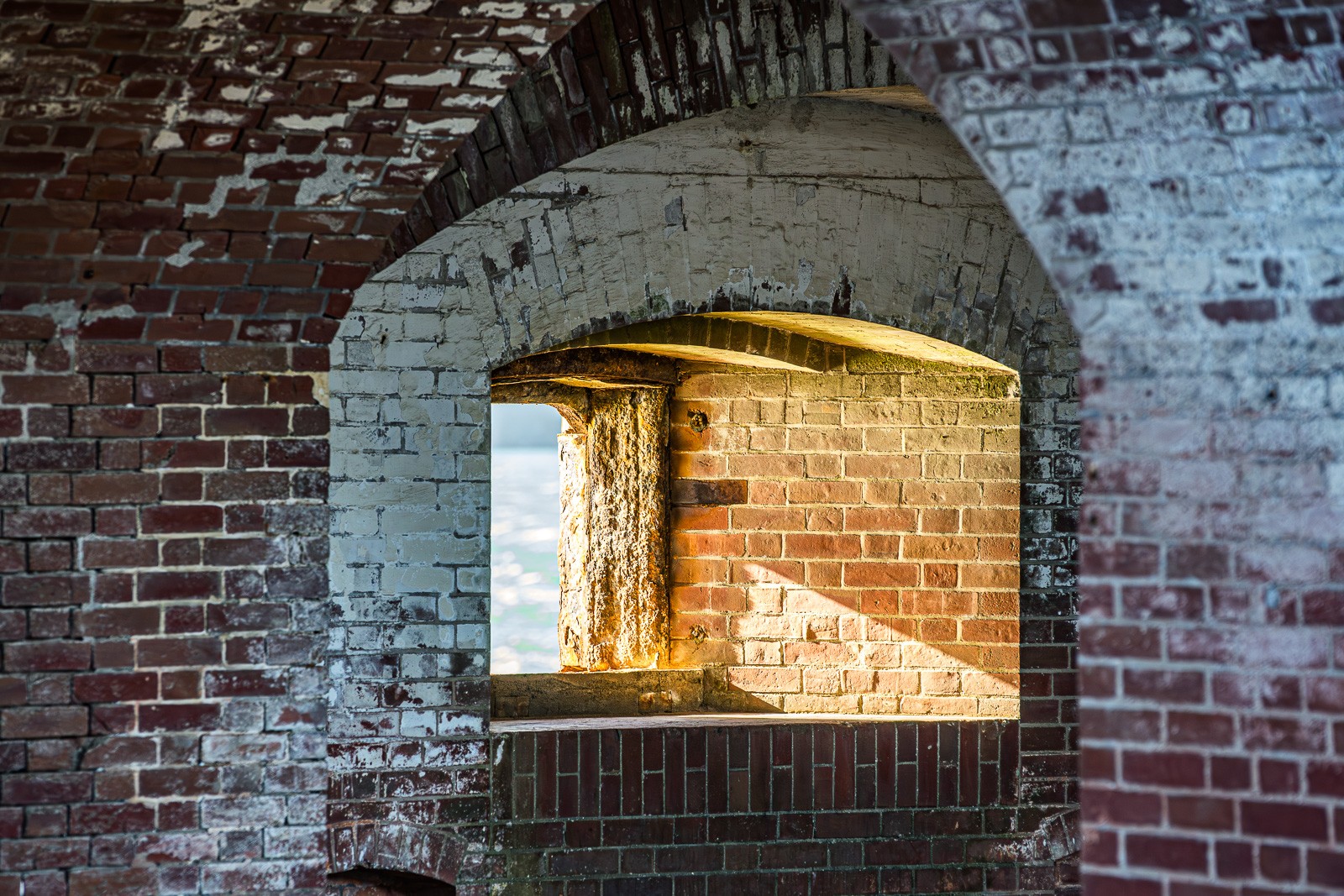 Shot from inside Fort Point. Golden sunlight pours through the window opening and falls in a warm diagonal across the brick wall to the right. The whole scene is about the light entering and hitting the aged masonry.