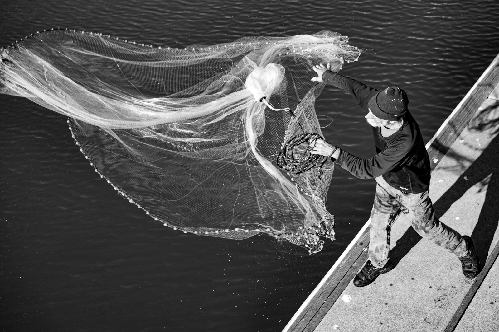 A fisherman photographed from above as he casts his net from a small boat, the circular net fully spread at the moment of release. A wedding gown hovering above dark water - until it dissolves in seconds.