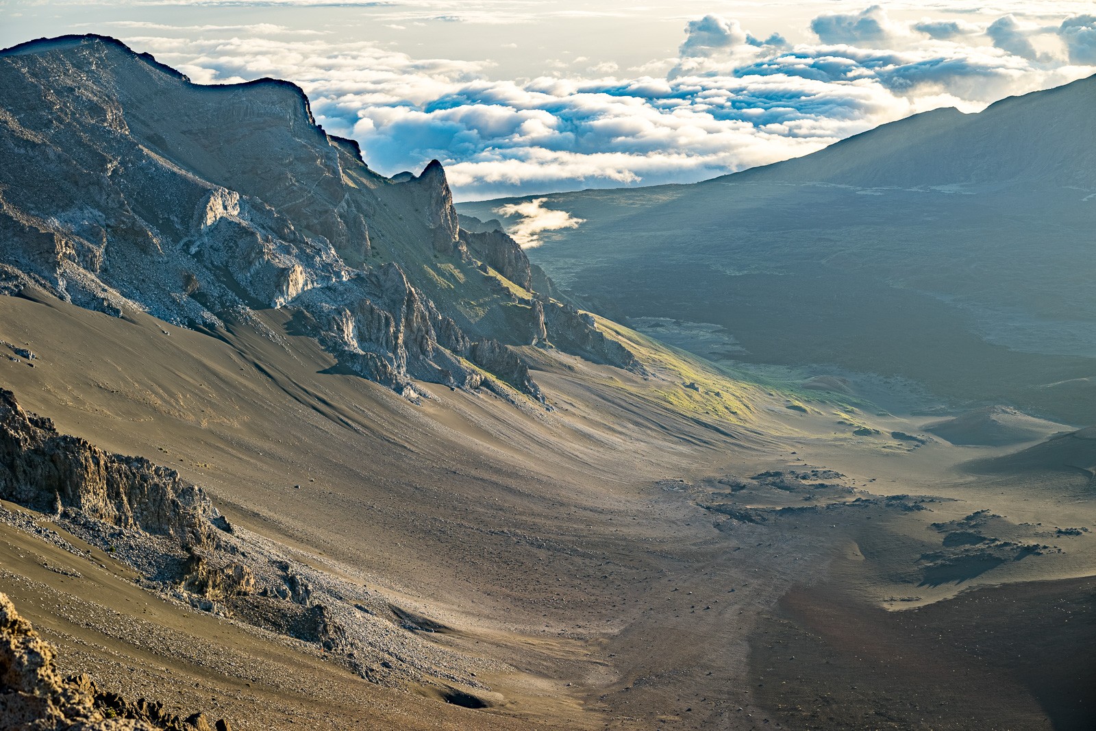 The barren upper slope of Haleakalā above the cloud line - cinder, rock, and nothing else. The crater rim visible on the horizon