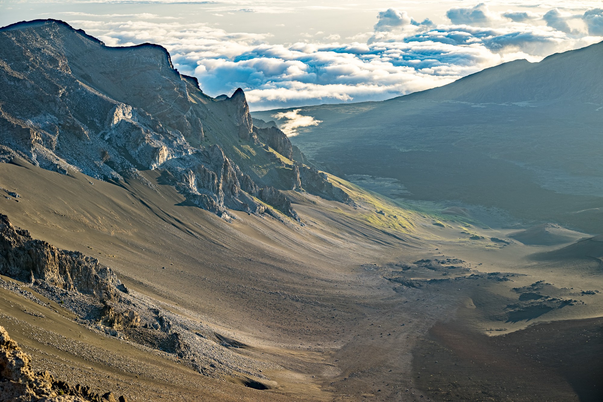The barren upper slope of Haleakalā above the cloud line - cinder, rock, and nothing else. The crater rim visible on the horizon