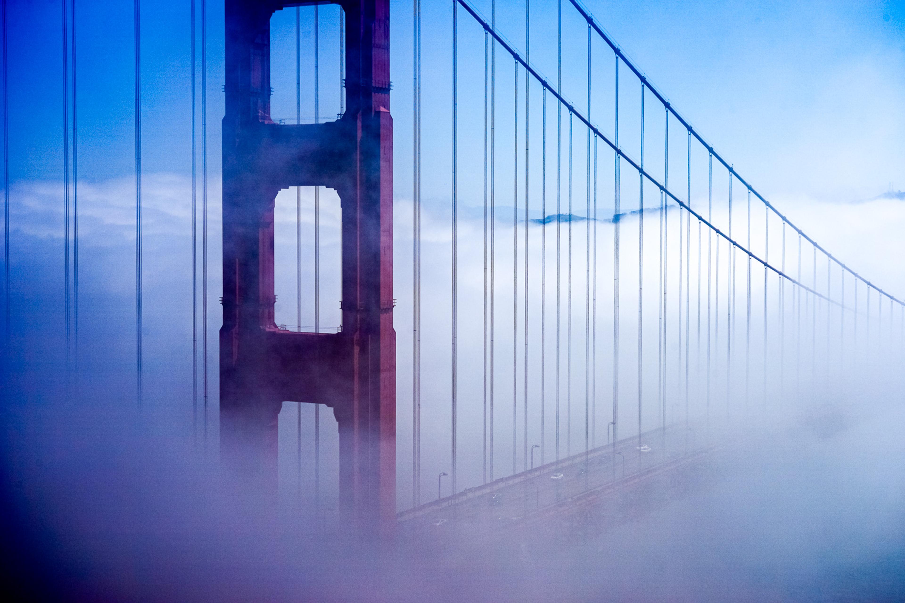 The Golden Gate Bridge north tower in heavy morning fog, the red paint barely visible through grey-white mist. The cables dissolve above