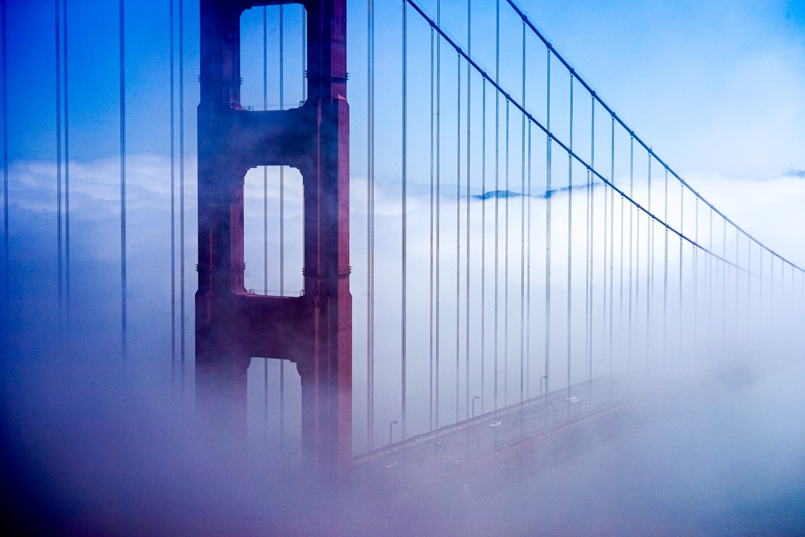 The Golden Gate Bridge north tower in heavy morning fog, the red paint barely visible through grey-white mist. The cables dissolve above