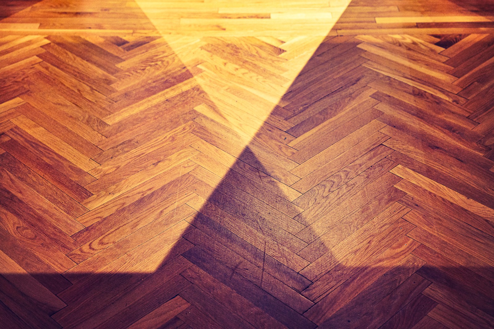 A herringbone wood floor photographed from directly above, the chevron pattern of the planks filling the frame entirely. Grain, joinery, and repeating geometry - texture treated as landscape.