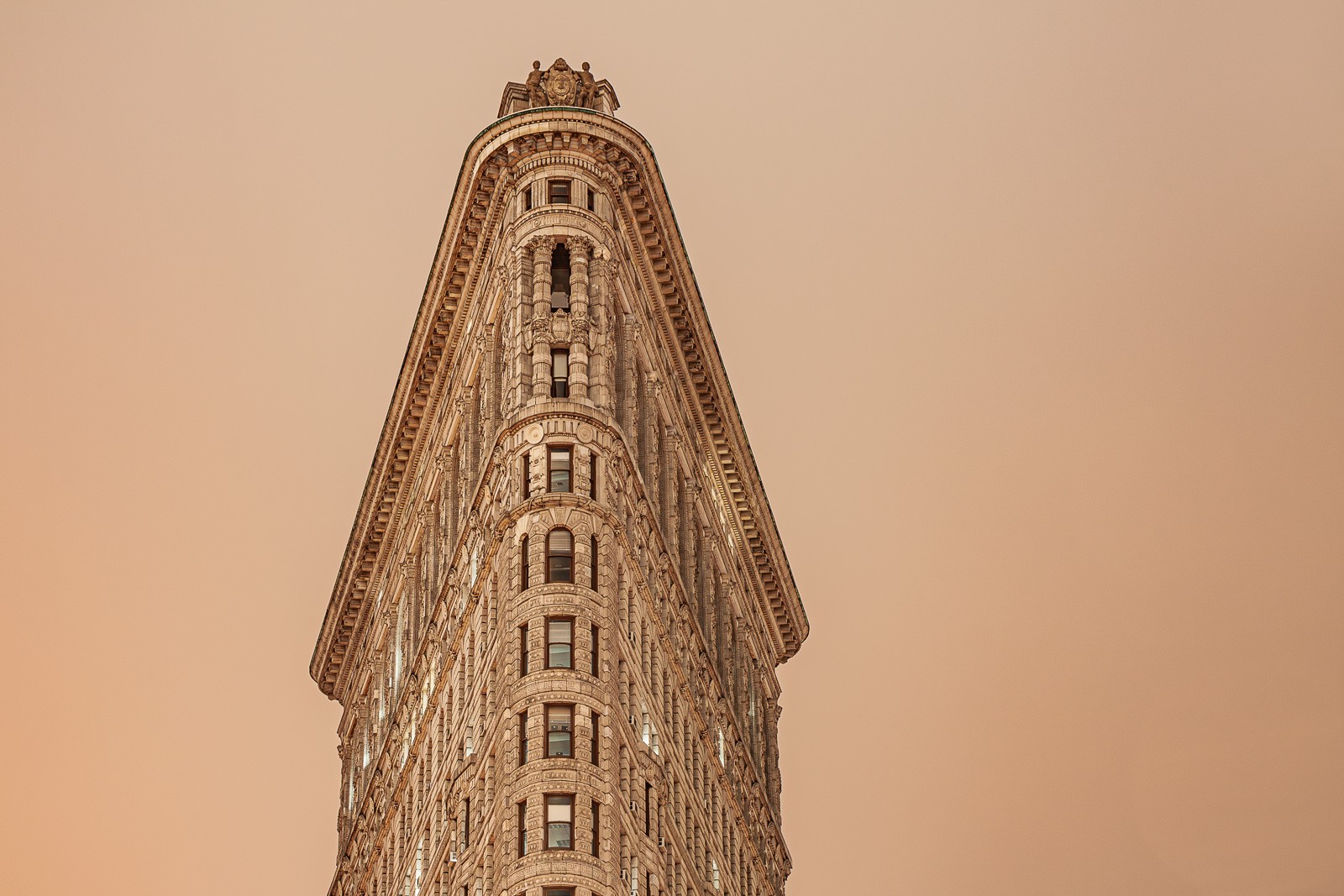 The Flatiron Building in warm dawn light, its triangular profile visible above the street-level trees. New York at the hour before the city fully wakes - the architecture clear, the light just arrived.