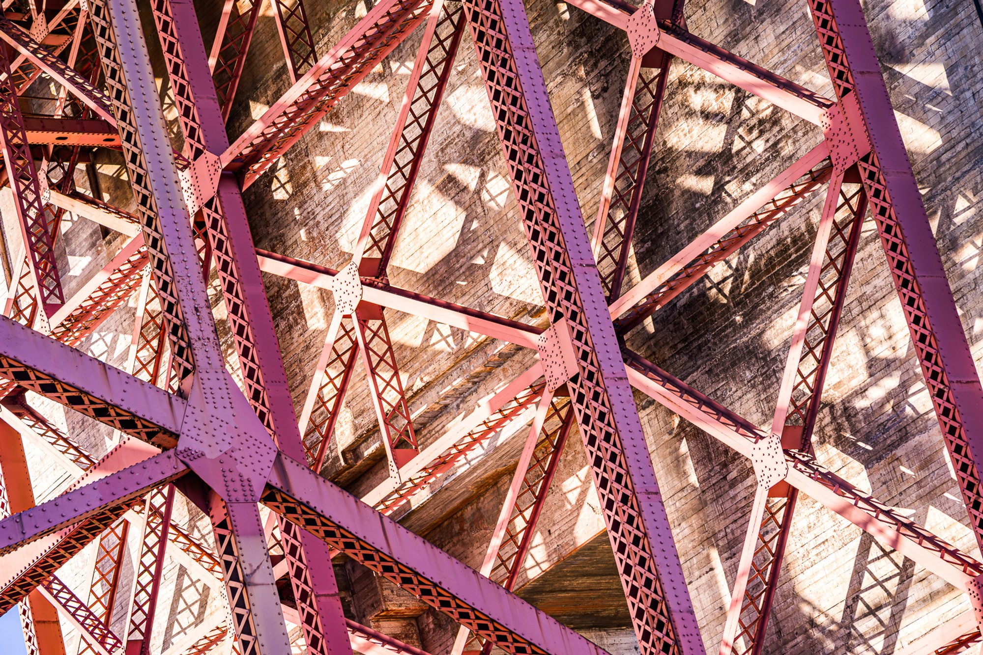 The structural underside of the Golden Gate Bridge photographed from within the span, the red-painted steel members and diagonal bracing filling the frame. The engineering that holds it up, seen from the inside for once.
