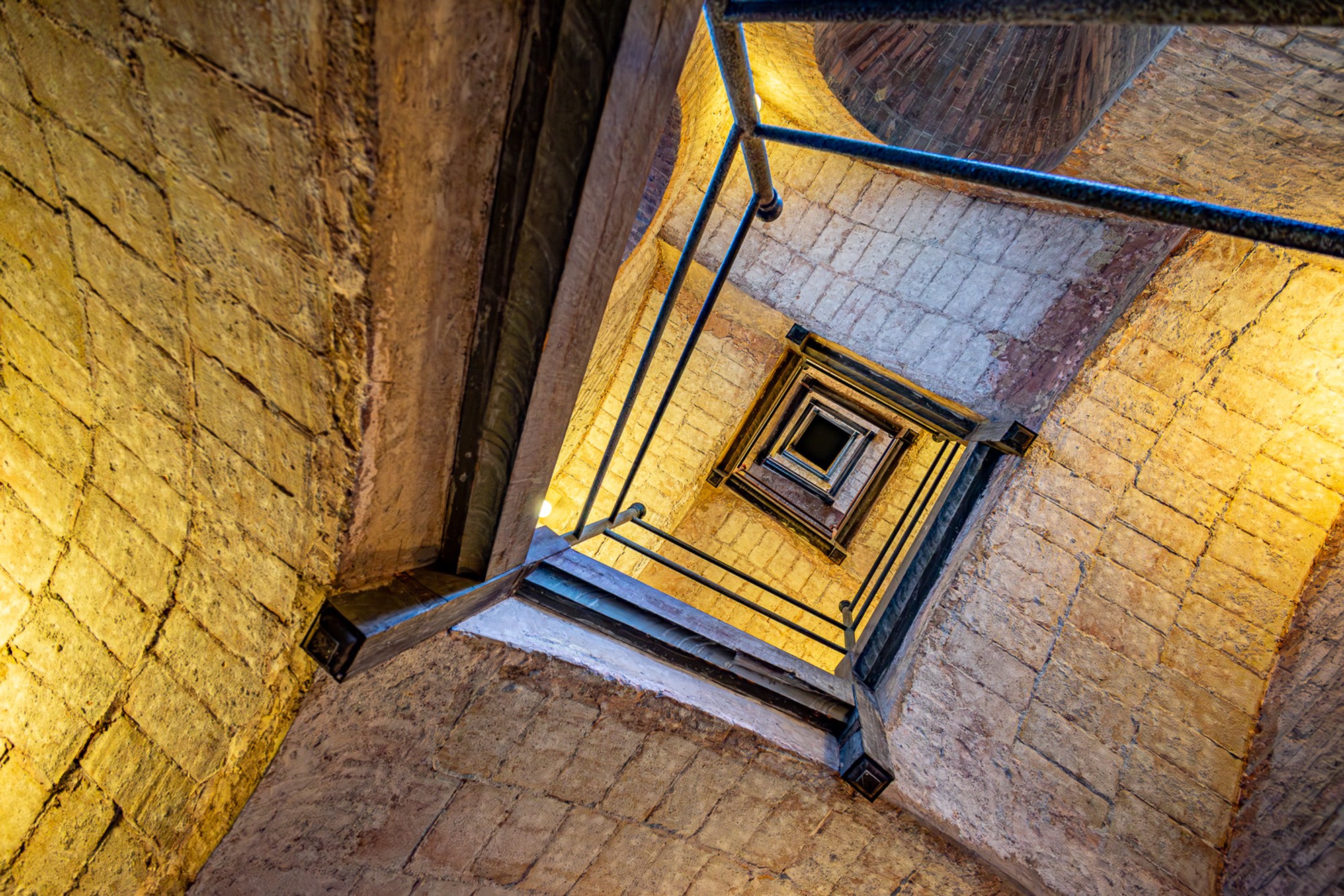 A stairwell in Barcelona shot from below, its amber-lit stone walls rising to a small skylight at the top. The walls form a square that narrows with distance - the geometry of the space revealed only from this exact viewpoint.