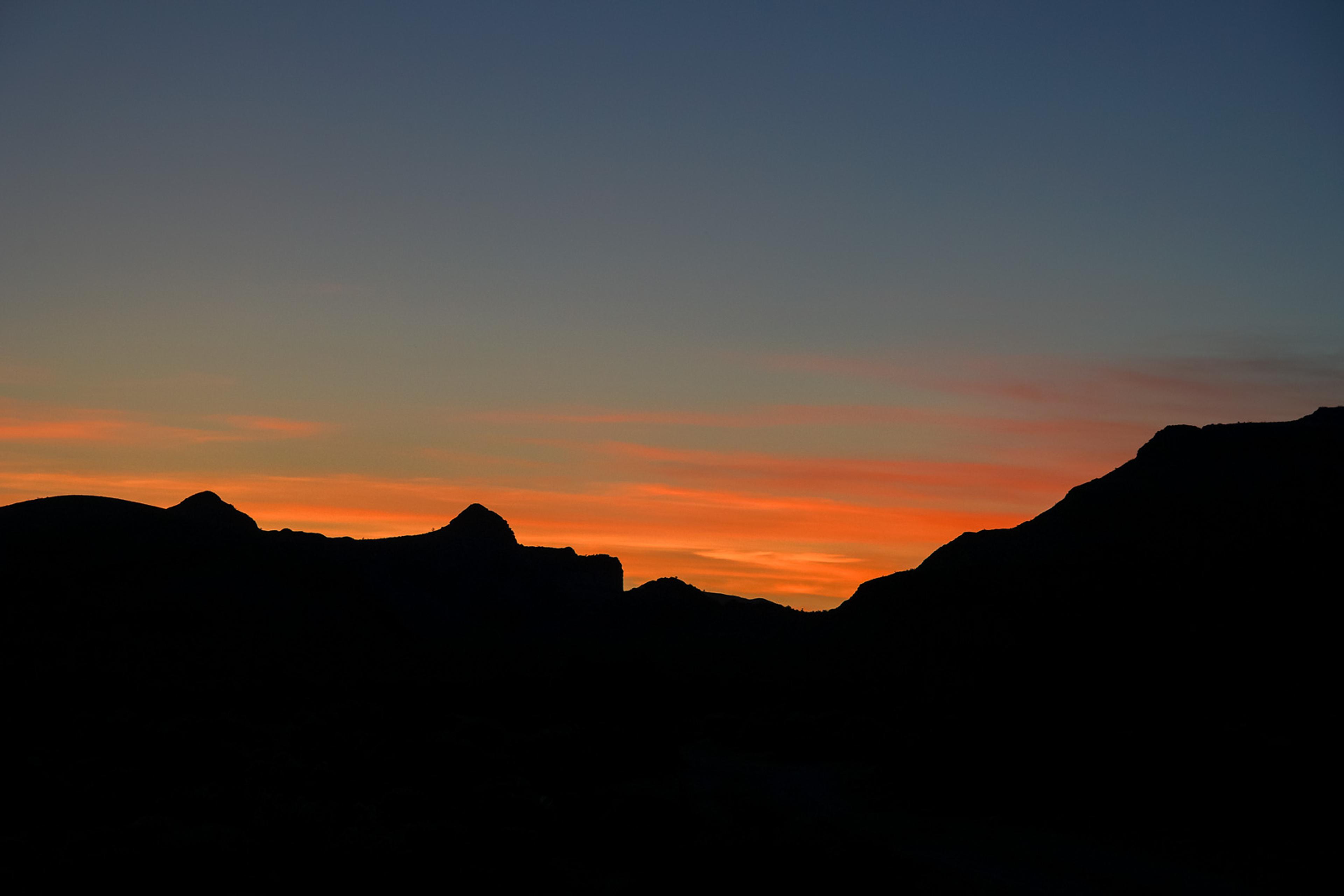 Dark mountain silhouettes stacked in layers against a Mojave sunrise, the horizon burning orange below deep blue. The sun is just below the ridgeline - the landscape holds everything in balance before the day begins.