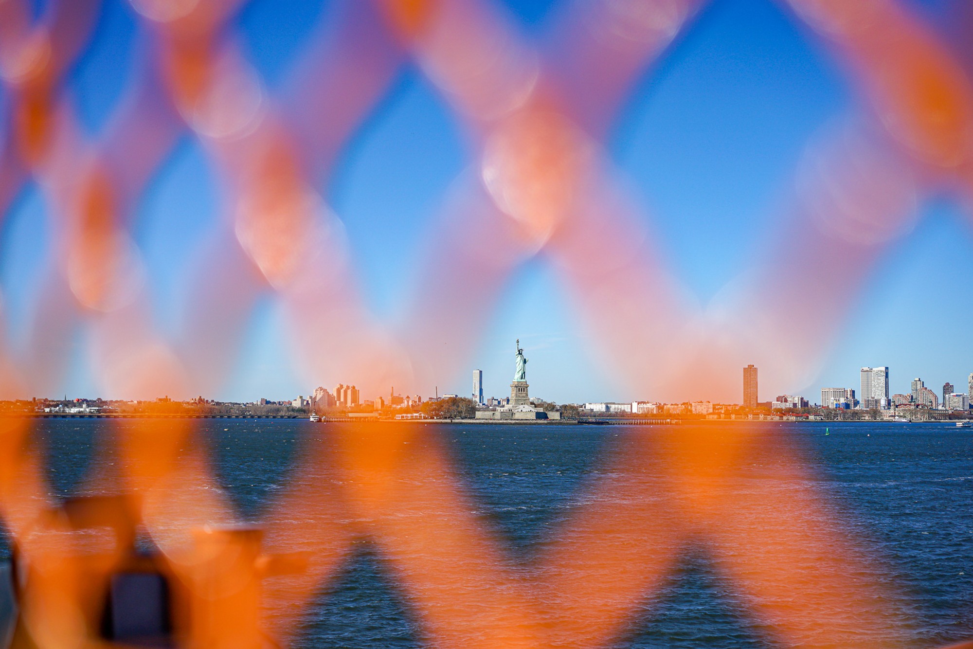 The Statue of Liberty seen through a chain-link fence, the fence sharp in the foreground while Liberty herself is soft and distant across the water. The orange diamond pattern occupies most of the frame - the icon reframed by what stands between.