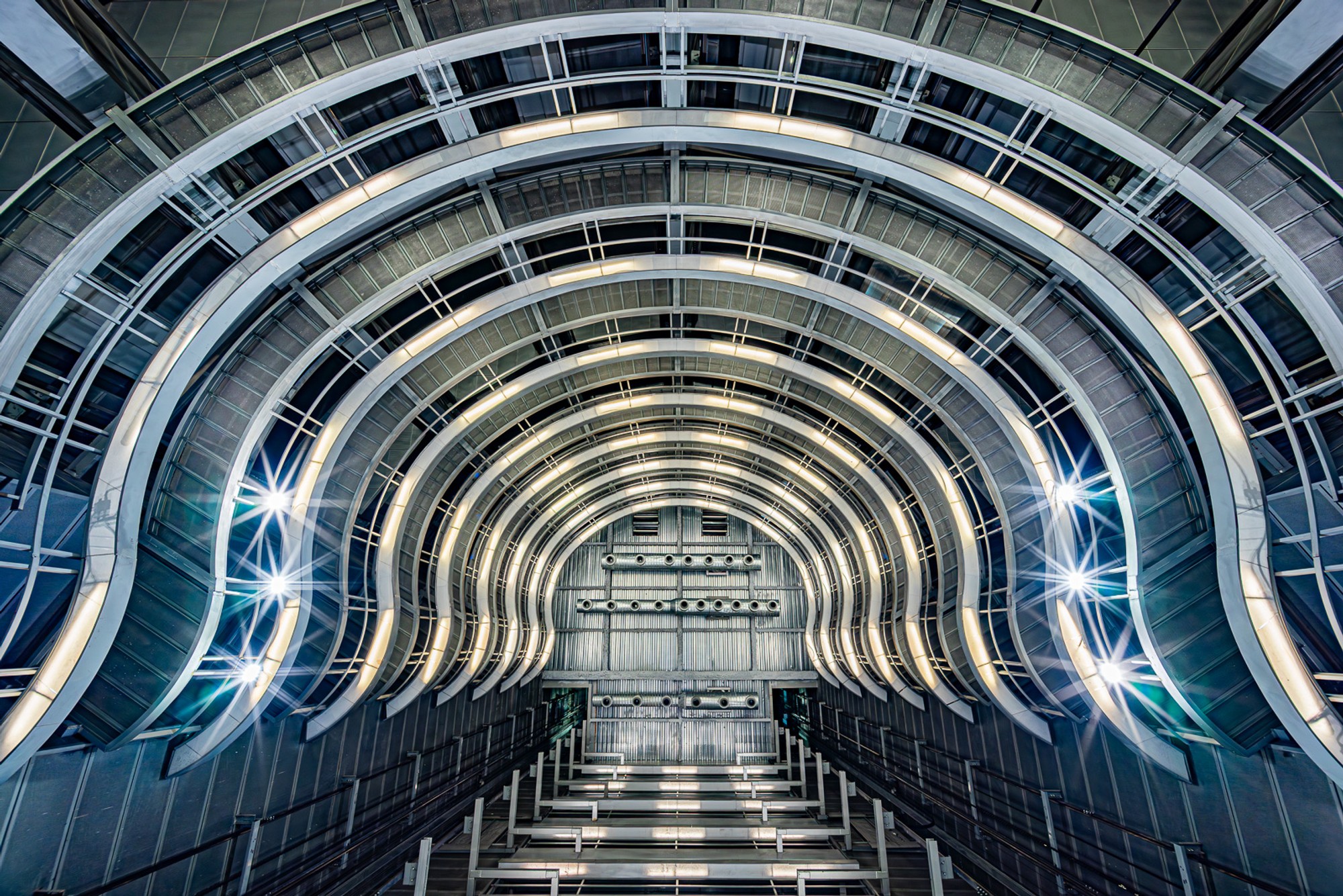 The vaulted ceiling of a Barcelona metro station, formed by interlocking steel arches that branch overhead in blue and silver. The structural engineering creates a tunnel that feels closer to cathedral than commute.