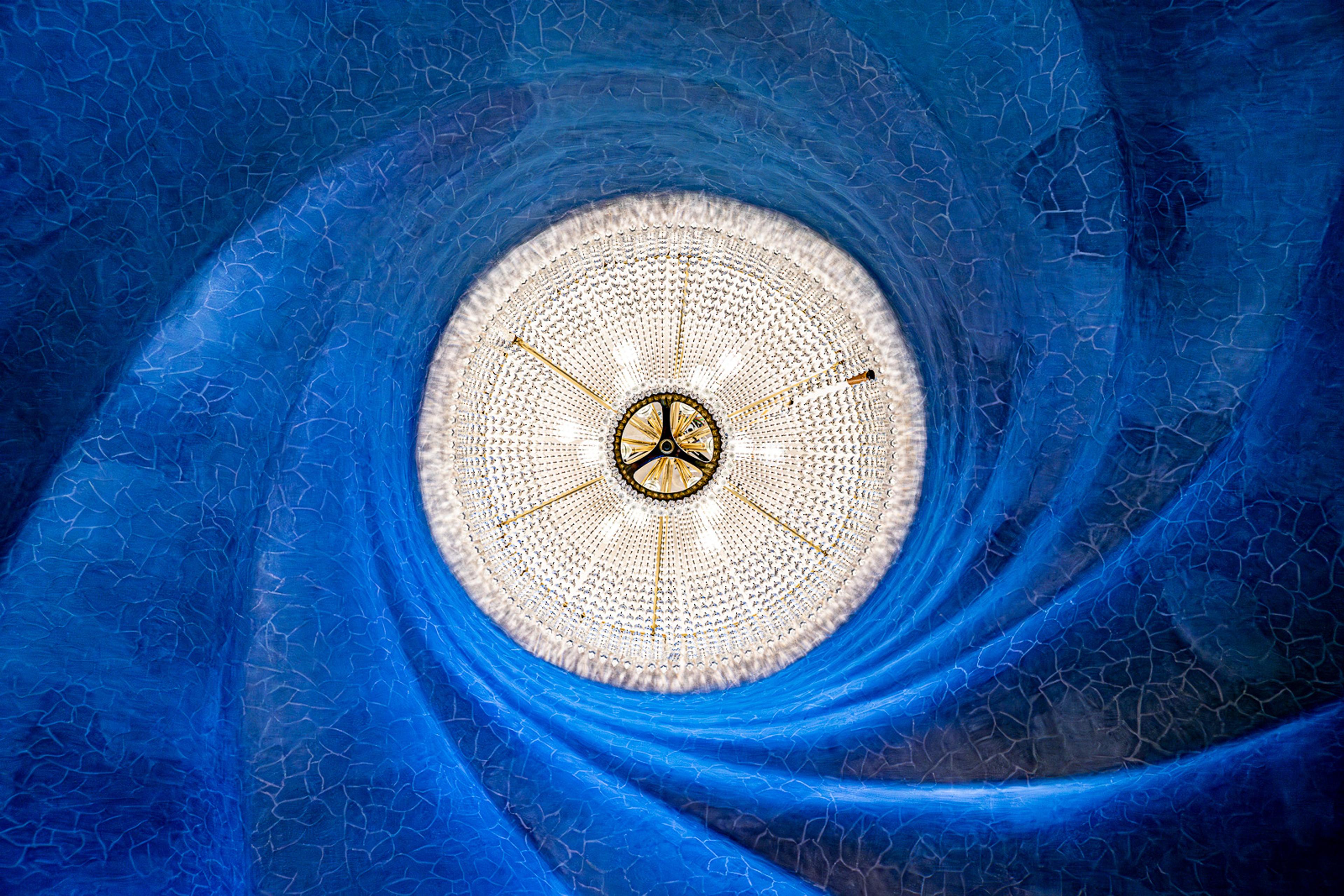 One of Gaudí's light installations inside the Casa Batlló, photographed from directly below. A circular disc of coloured glass and metalwork glows in blue and gold - the same structural logic as the columns outside, applied at the smallest possible scale.