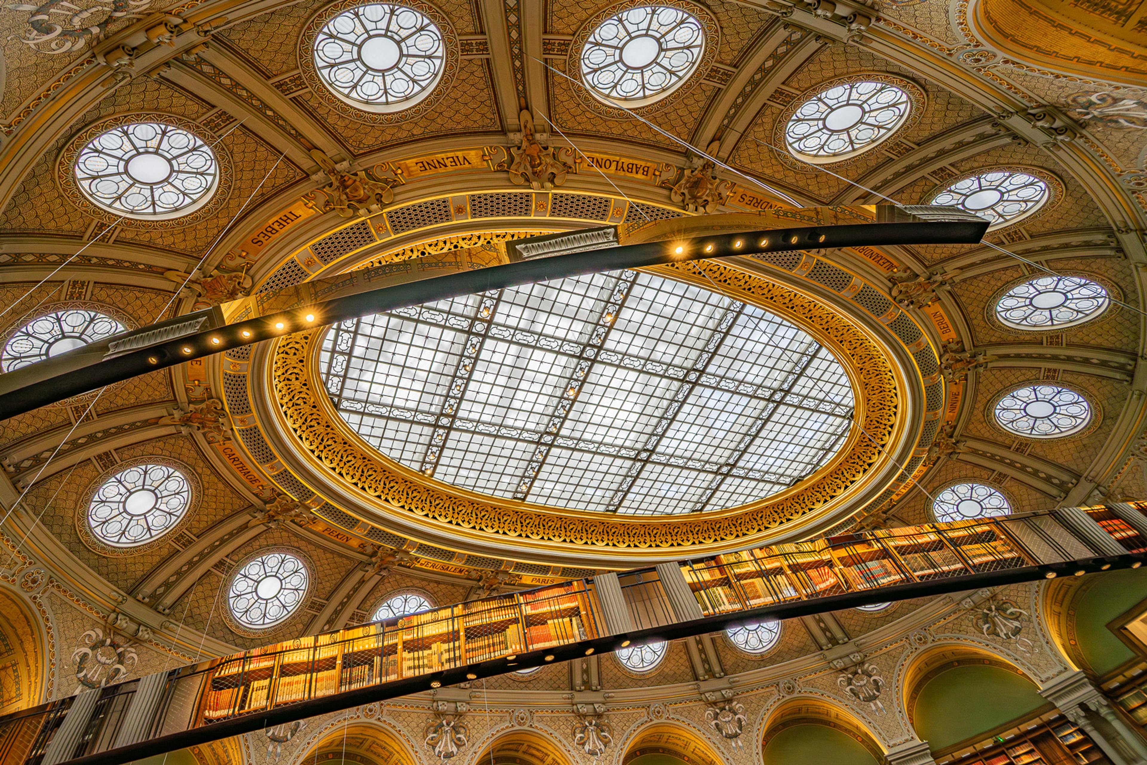 The barrel-vaulted reading room of a grand Parisian library, green-shaded lamps running the full length of the hall. A space designed for concentration and beauty simultaneously - the kind that cities build once and maintain forever.