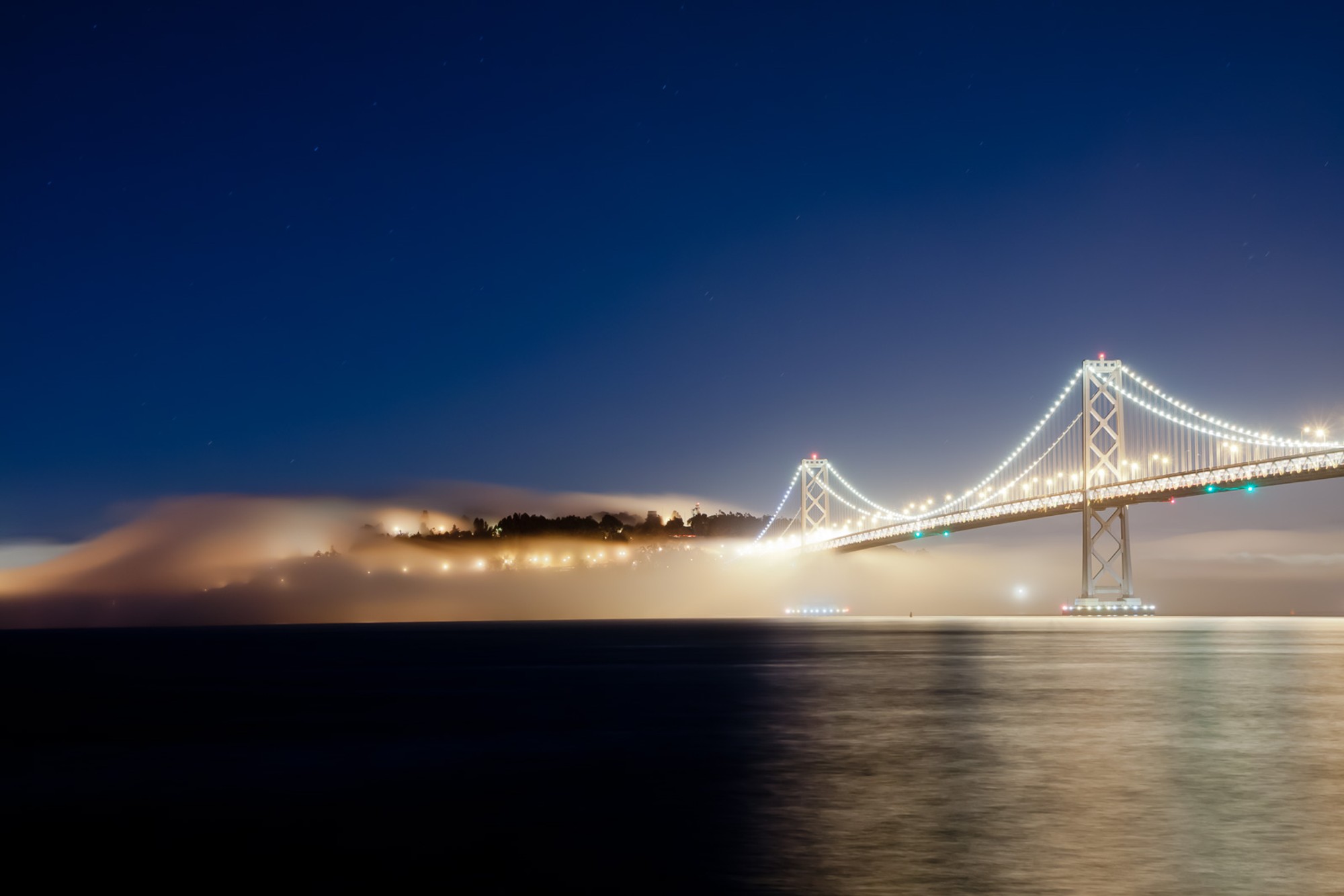 A long exposure of the Bay Bridge at night, the light trails of passing traffic arcing across the span above still black water. The bridge becomes light