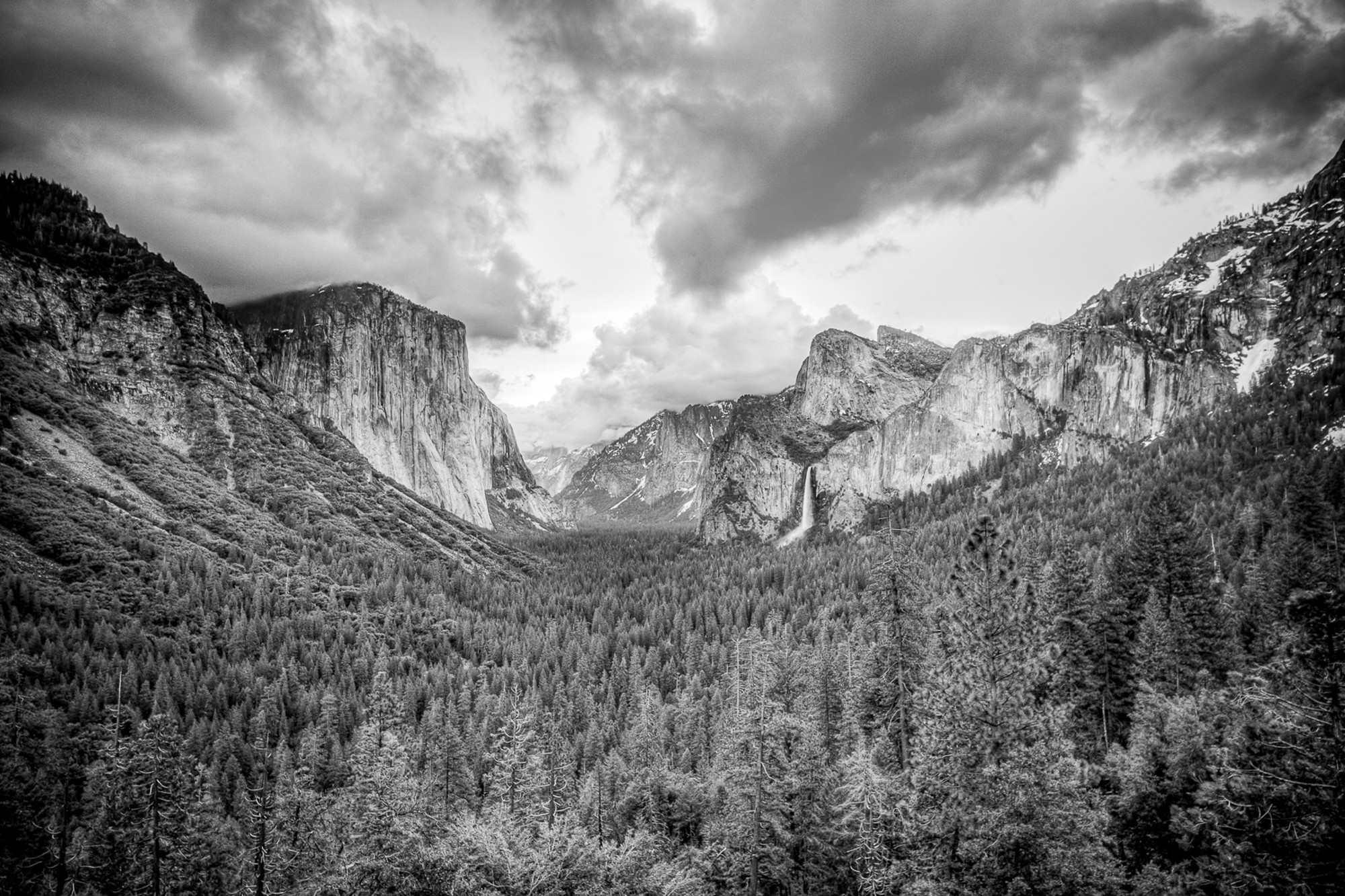 The classic Yosemite Valley panorama in black and white - El Capitan on the left, Bridalveil Fall on the right, Half Dome in the distance. Dark storm clouds above the valley walls give the scene a weight the colour version rarely achieves.