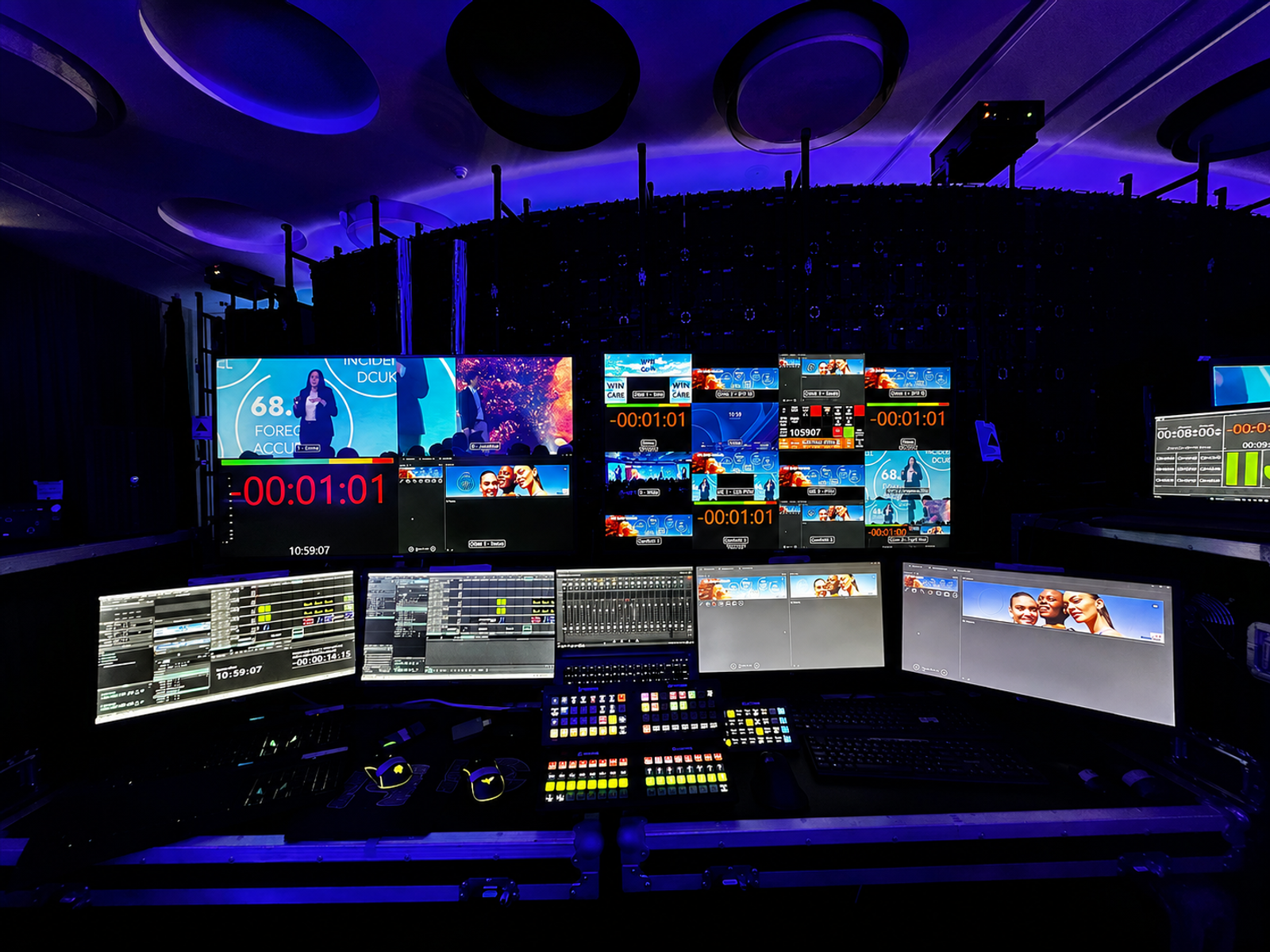 Professional event production control room featuring multiple video screens, live broadcast feeds and technical equipment illuminated with blue and purple lighting