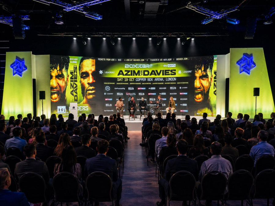 A professionally produced conference with a seated audience facing a large, brightly lit stage featuring a panel discussion and bold digital event graphics.
