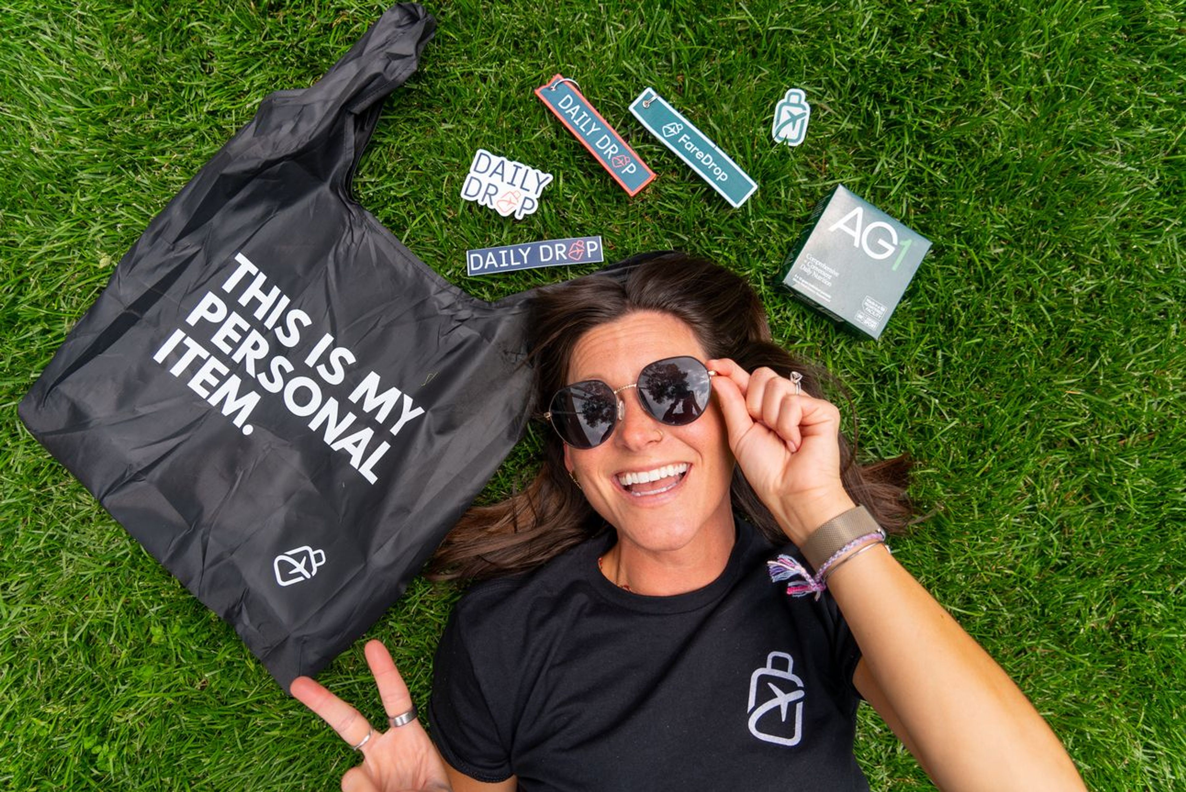 Kara lying on the grass with FareDrop sitckers, bag tags, a tote, and AG1