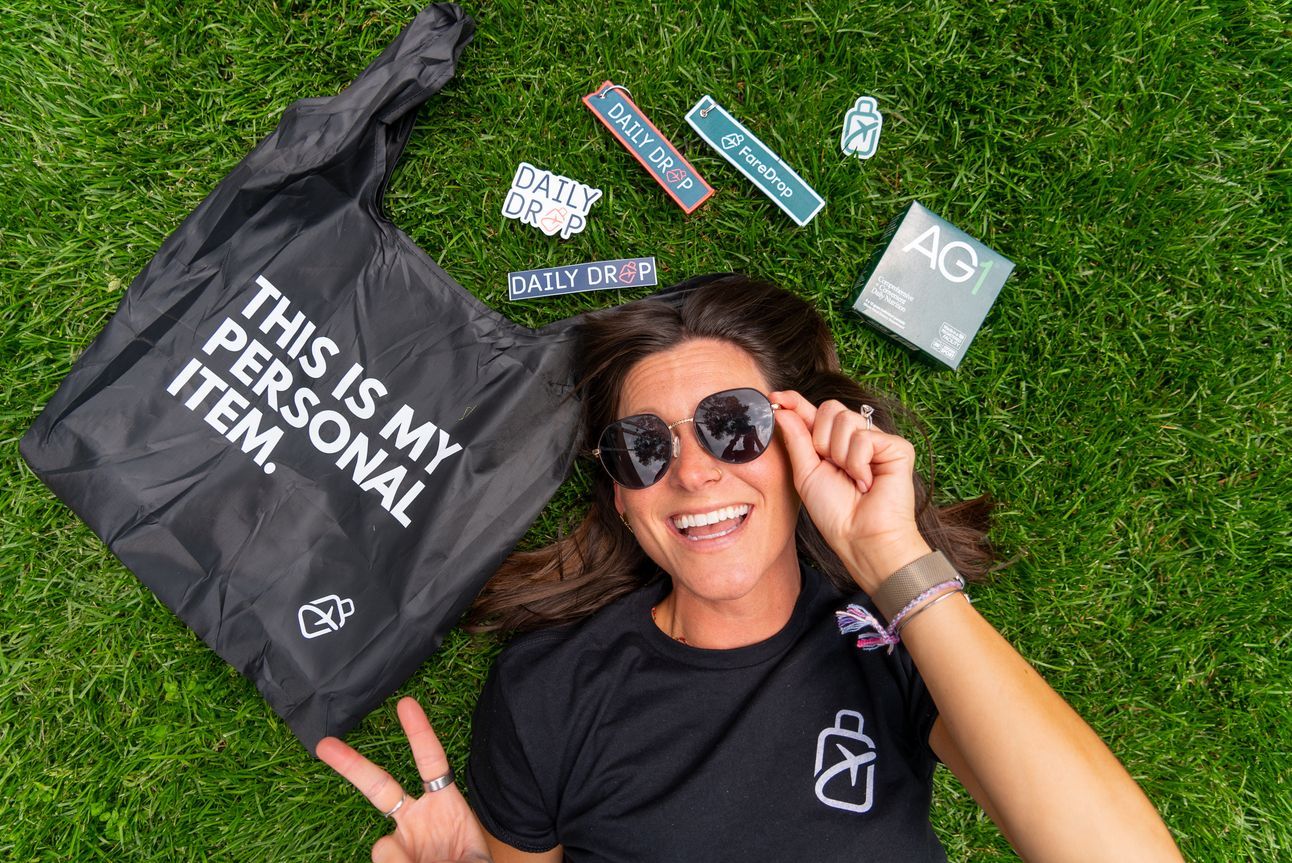 Kara lying on the grass with FareDrop sitckers, bag tags, a tote, and AG1