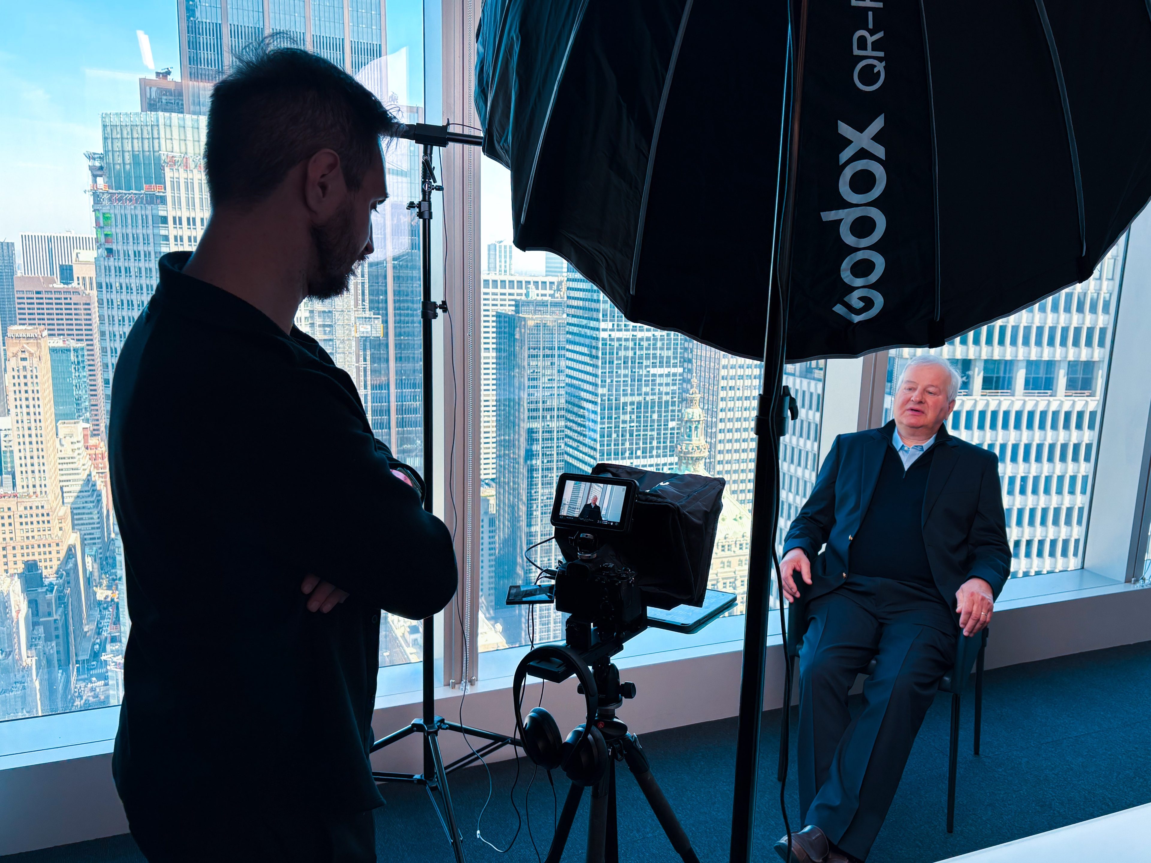 Director beside the camera during a seated interview, Midtown skyline through the windows