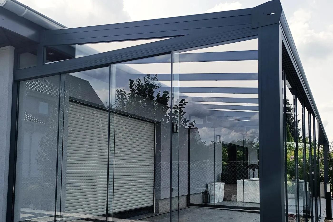 Überdachung in Anthrazit mit Glasschiebewänden an einem hellen Haus, an der Seite ein gläserner Spitzkeil unter dem geneigten Dach