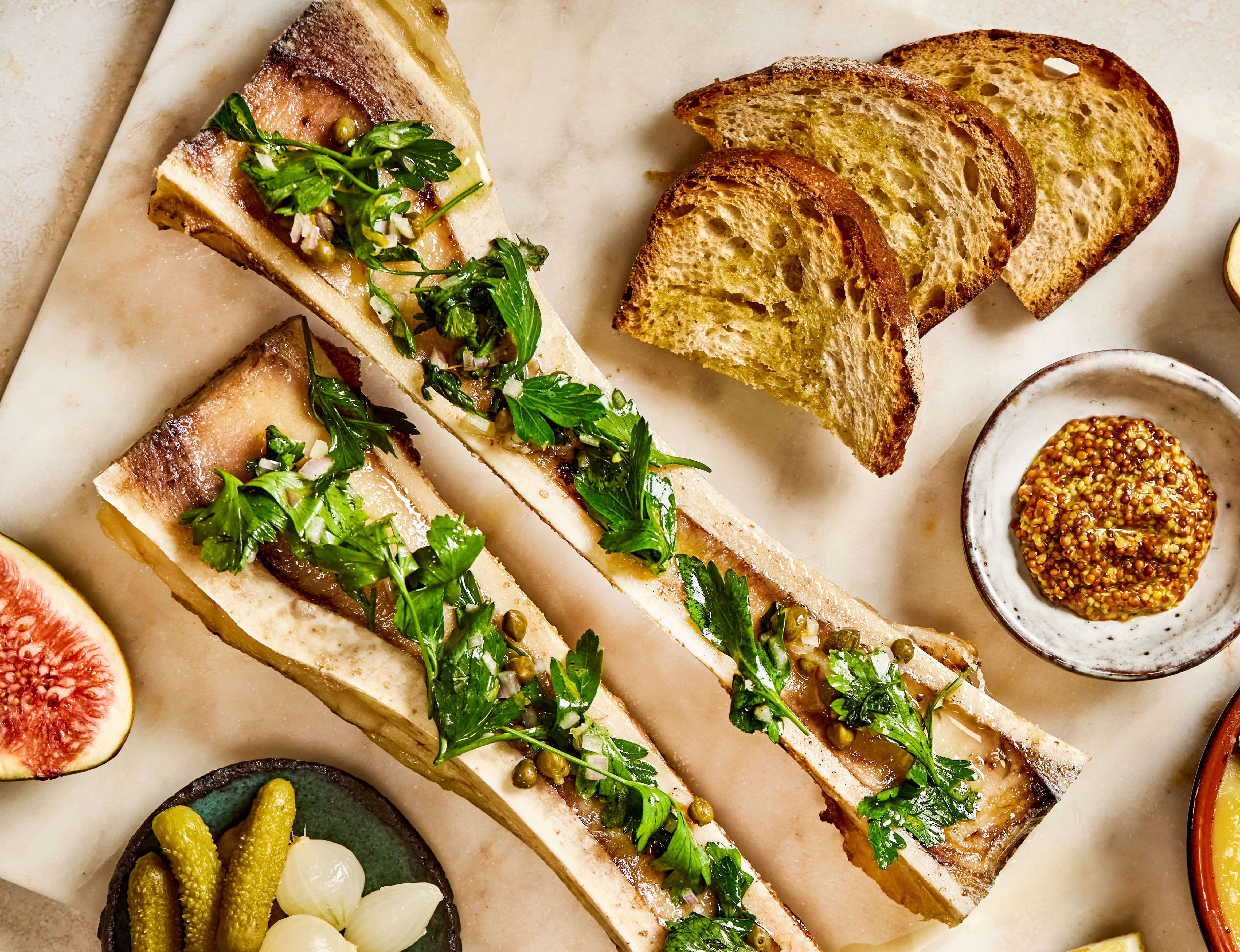 Gratinert margbein og kyllingleverterrine på et bord, skåler med toast, pickles, sennep og fersk fiken.