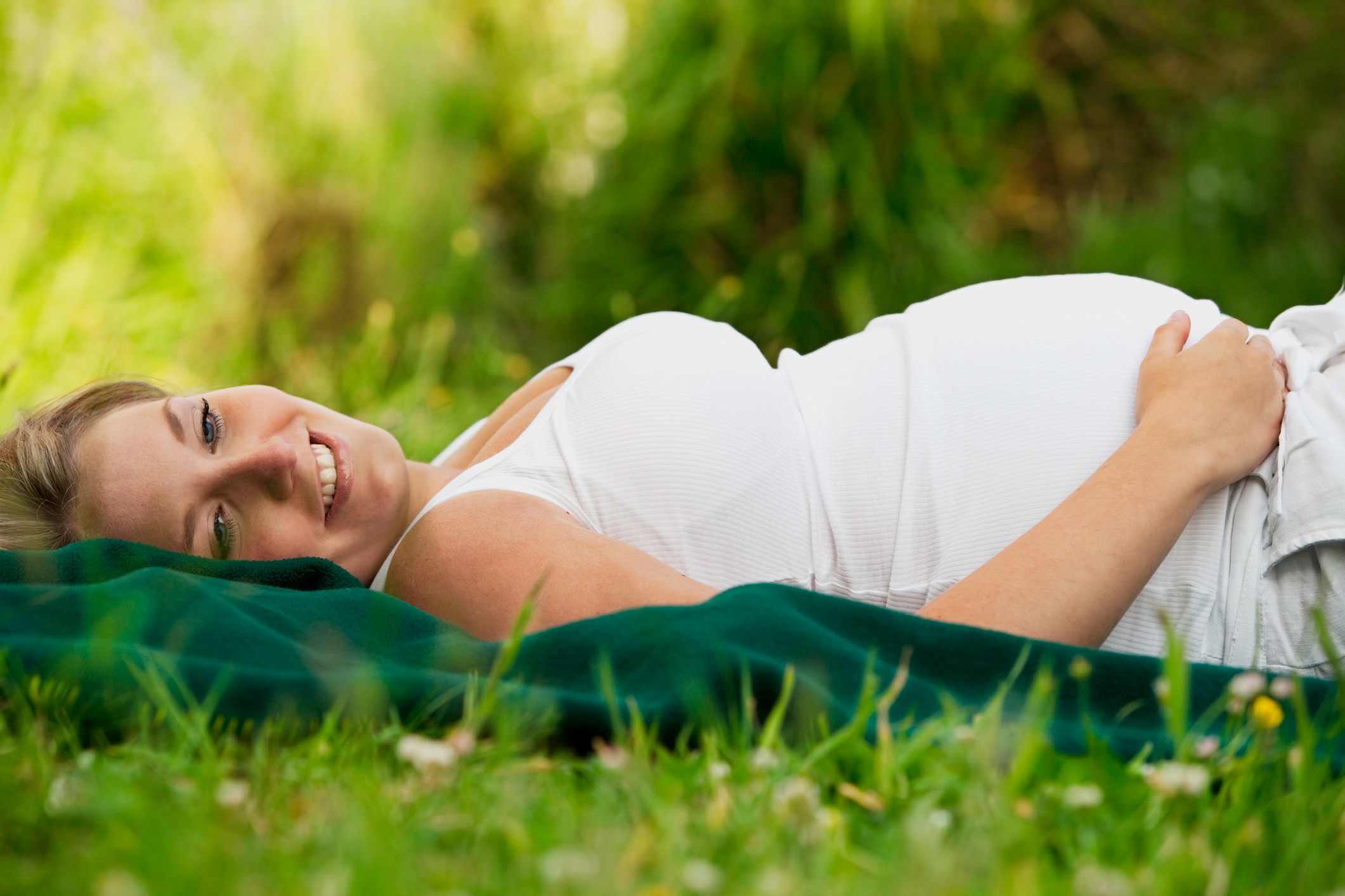 Gravid dame i hvit topp som ligger på en gressplen og smiler.