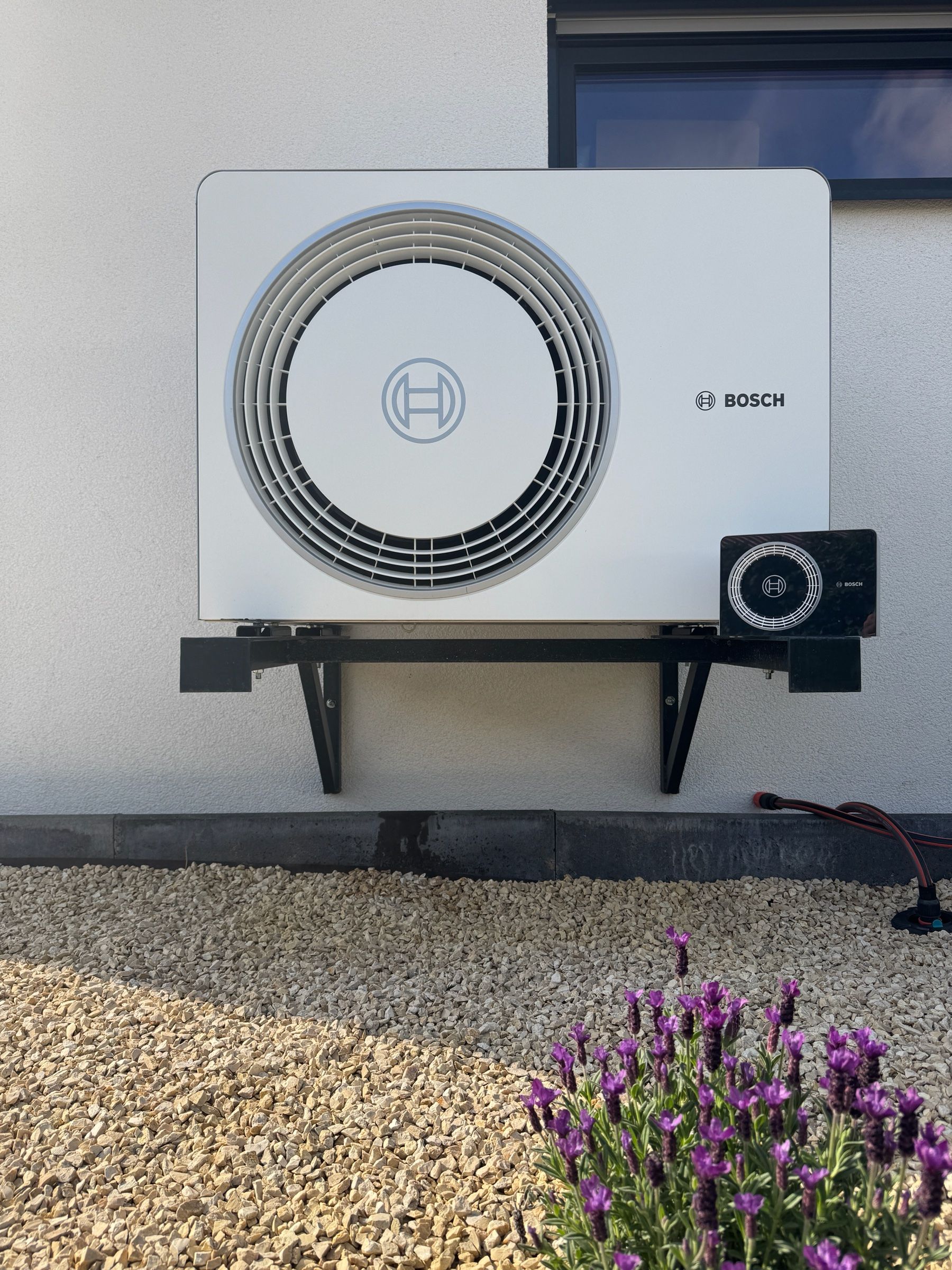Außeneinheit einer Bosch-Wärmepumpe auf einer Wandkonsole über einem Kiesbeet mit Lavendel