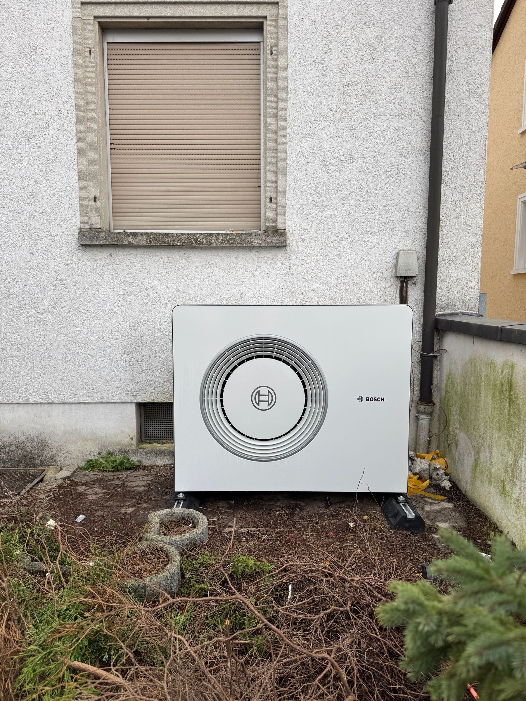 Außeneinheit einer Bosch-Wärmepumpe vor einer weiß verputzten Hauswand unter einem Fenster mit Rollladen