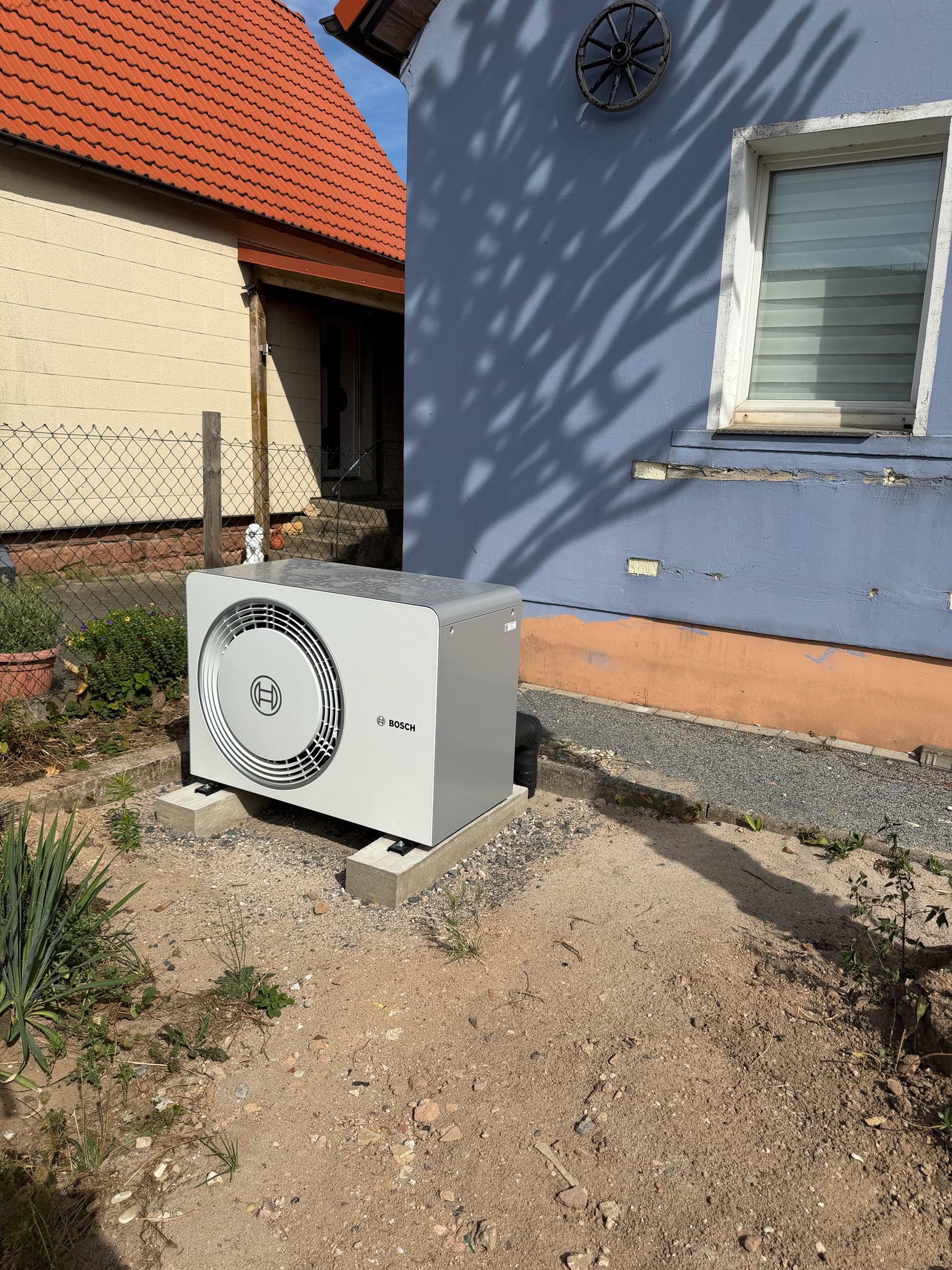 Außeneinheit einer Bosch-Wärmepumpe auf einem Sockel im Garten neben einem Wohnhaus