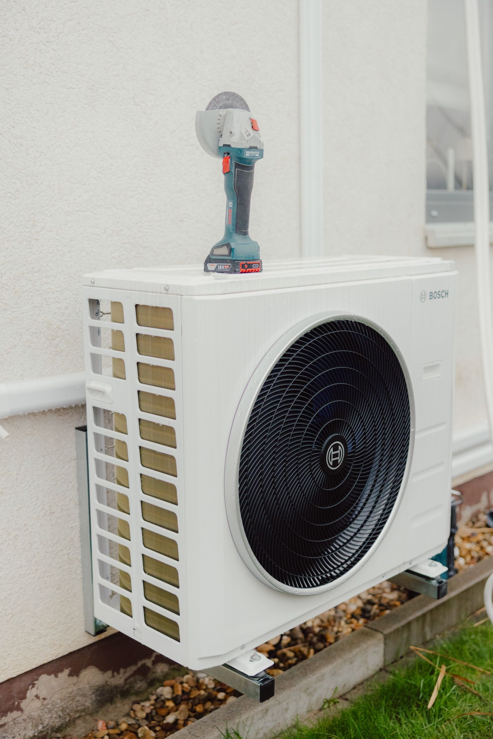 Außengerät einer Bosch-Klimaanlage auf einer Wandkonsole an einer hellen Hauswand, obenauf liegt ein Akku-Winkelschleifer