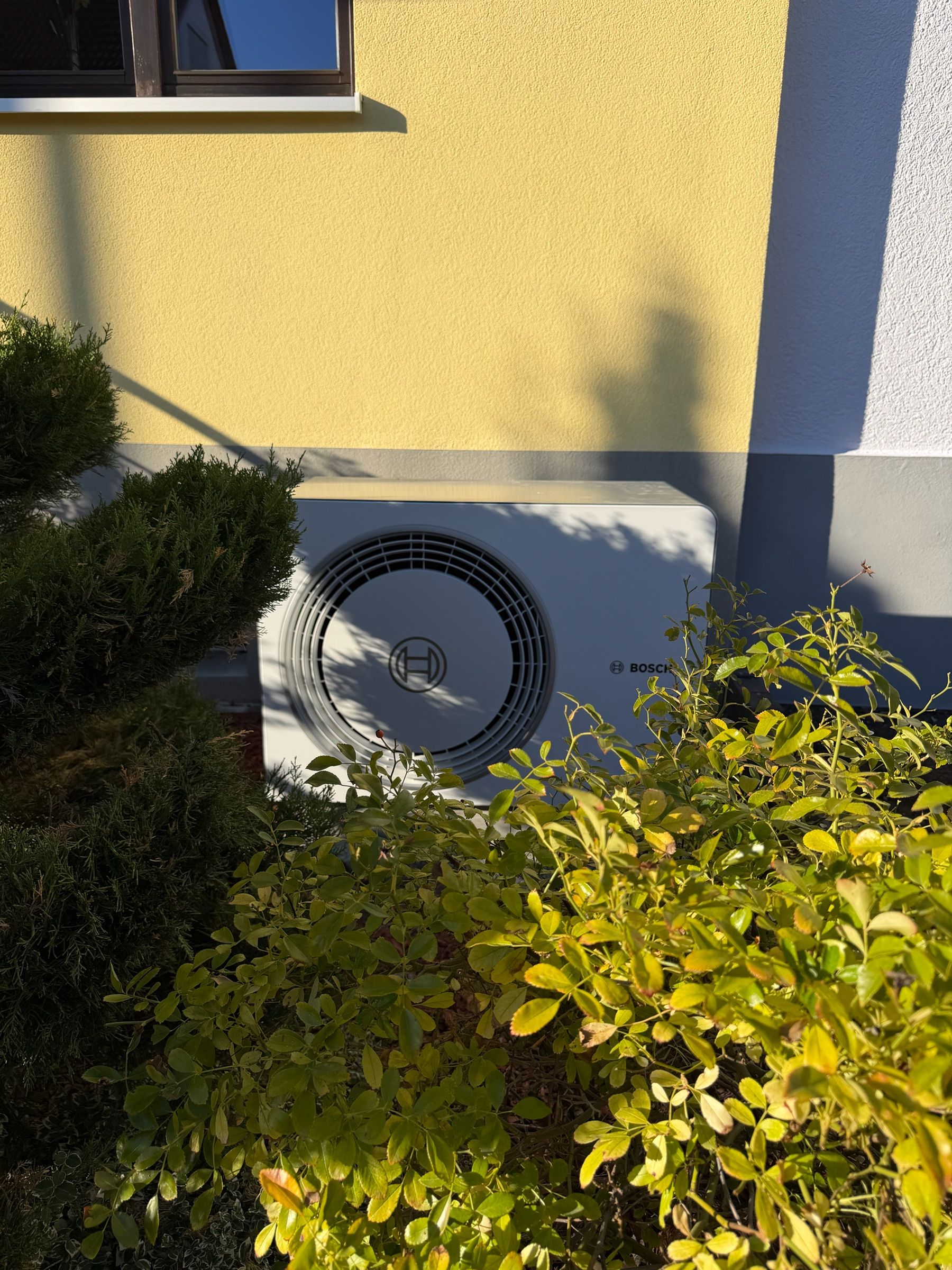 Außeneinheit einer Bosch-Wärmepumpe vor einer gelben Hauswand, teilweise hinter Sträuchern