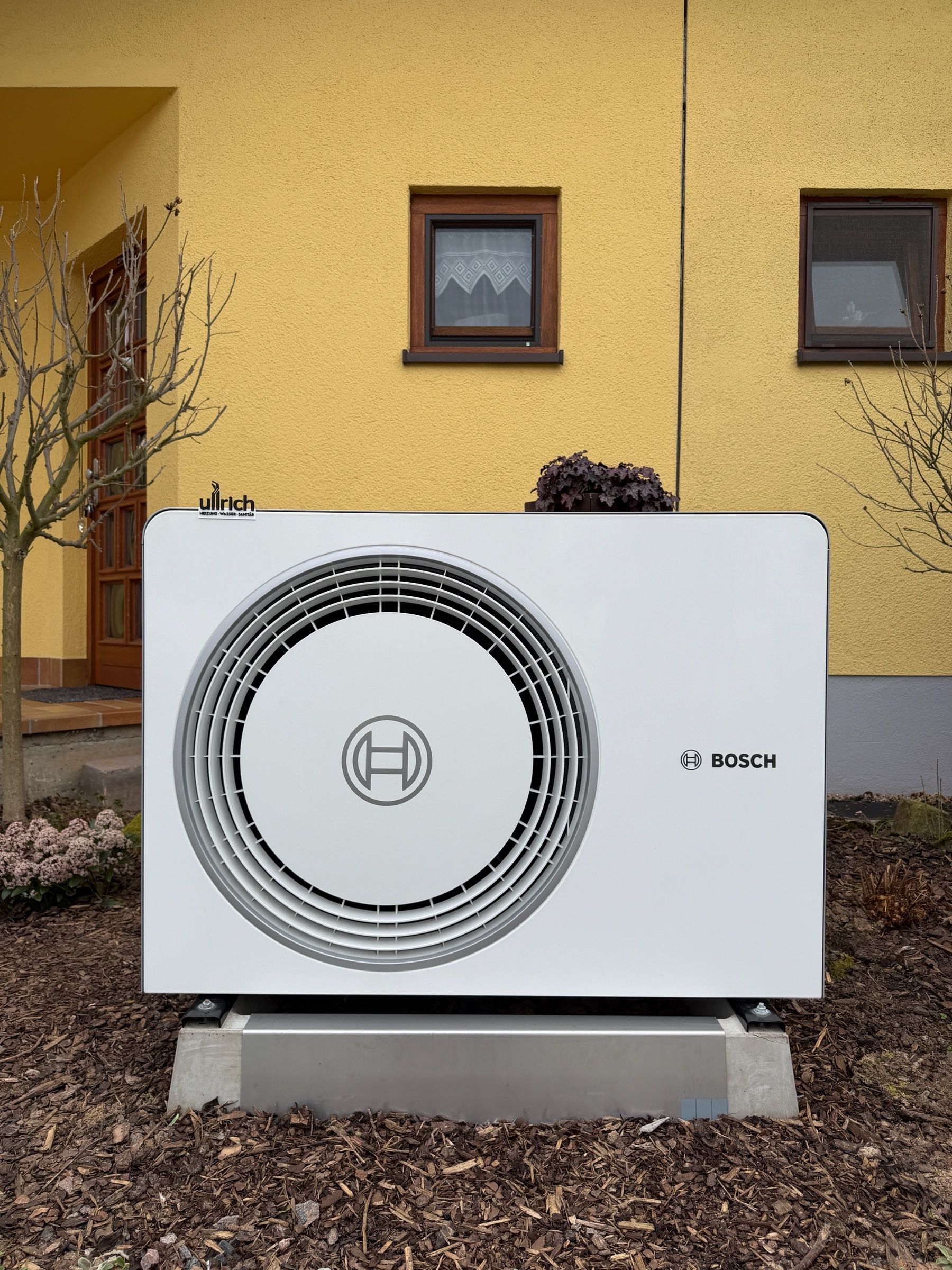 Außeneinheit einer Bosch-Wärmepumpe mit Ullrich-Aufkleber vor einer gelben Hauswand