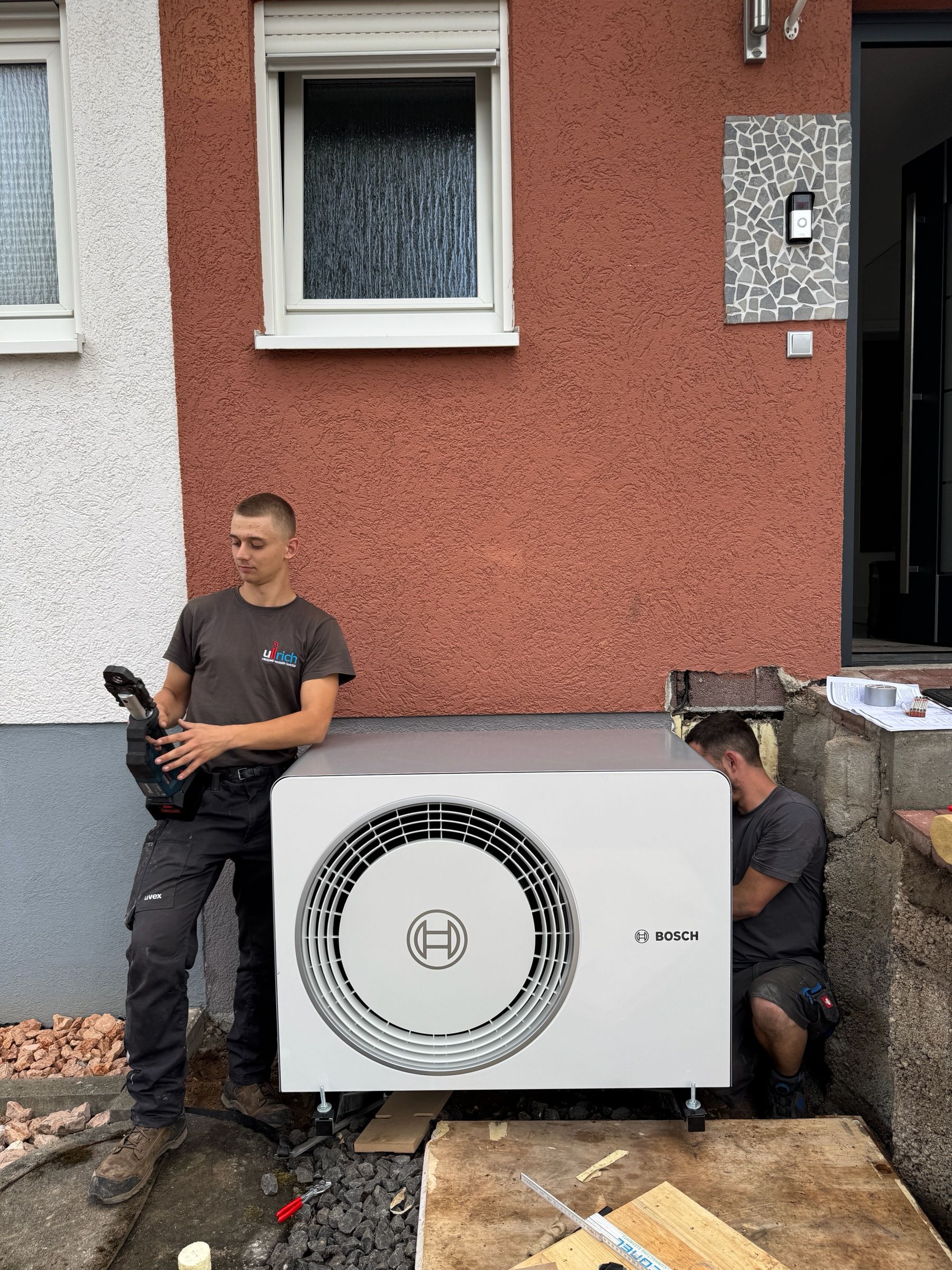 Monteure bei der Aufstellung der Außeneinheit einer Bosch-Wärmepumpe vor einer roten Hauswand