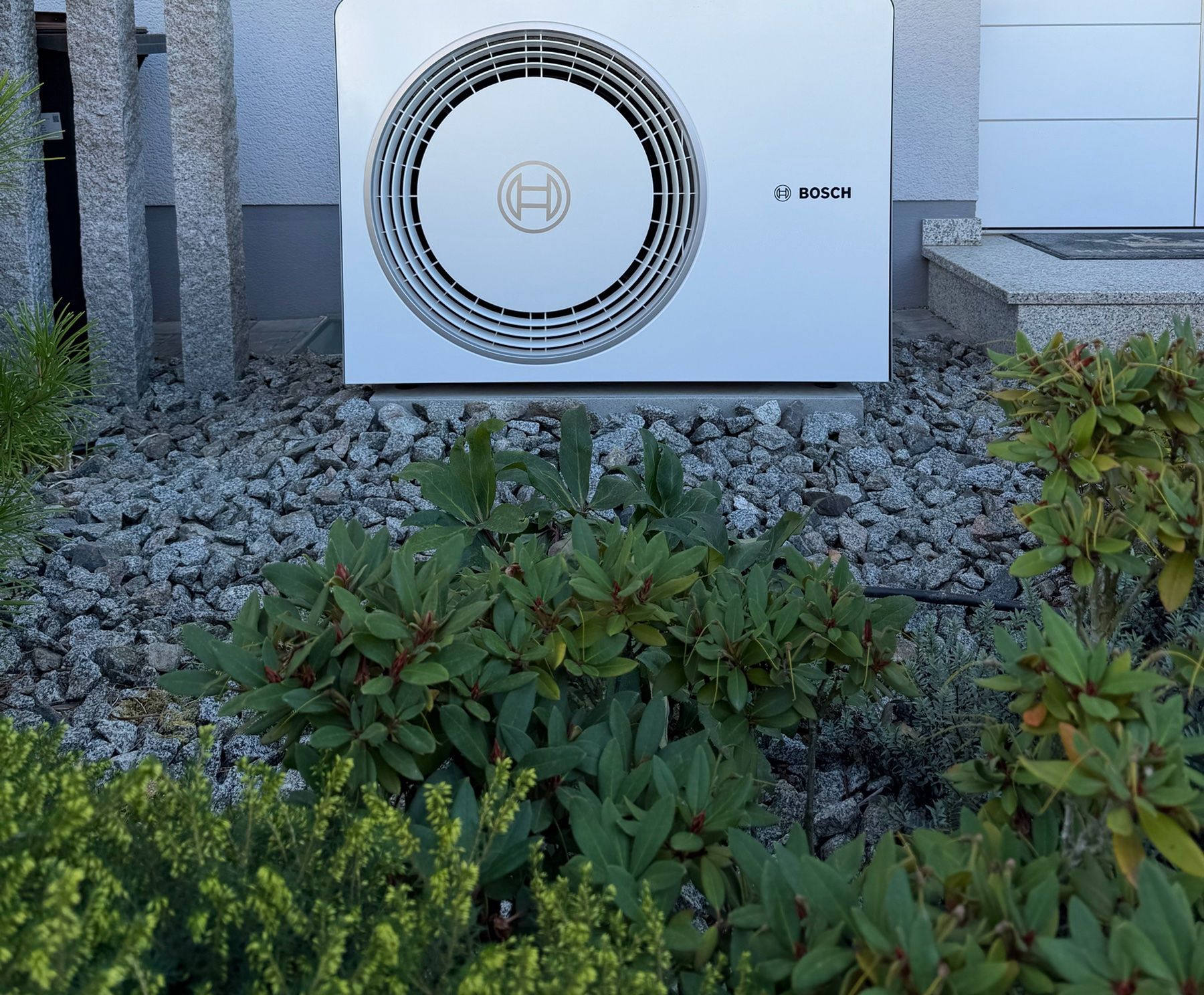 Außeneinheit einer Bosch-Wärmepumpe in einem Kiesbeet mit Sträuchern