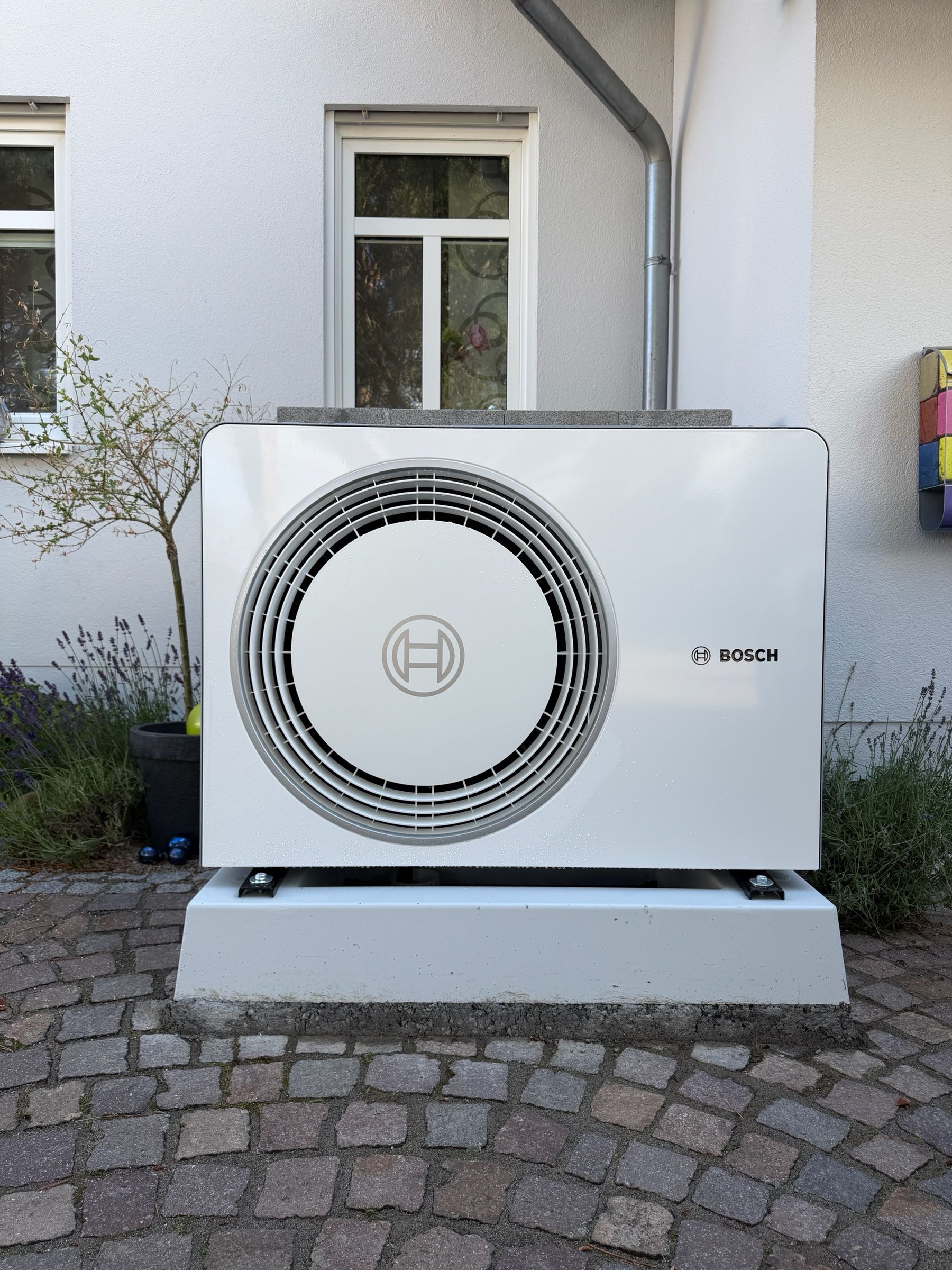 Außeneinheit einer Bosch-Wärmepumpe auf einem hellen Sockel in einem gepflasterten Hof