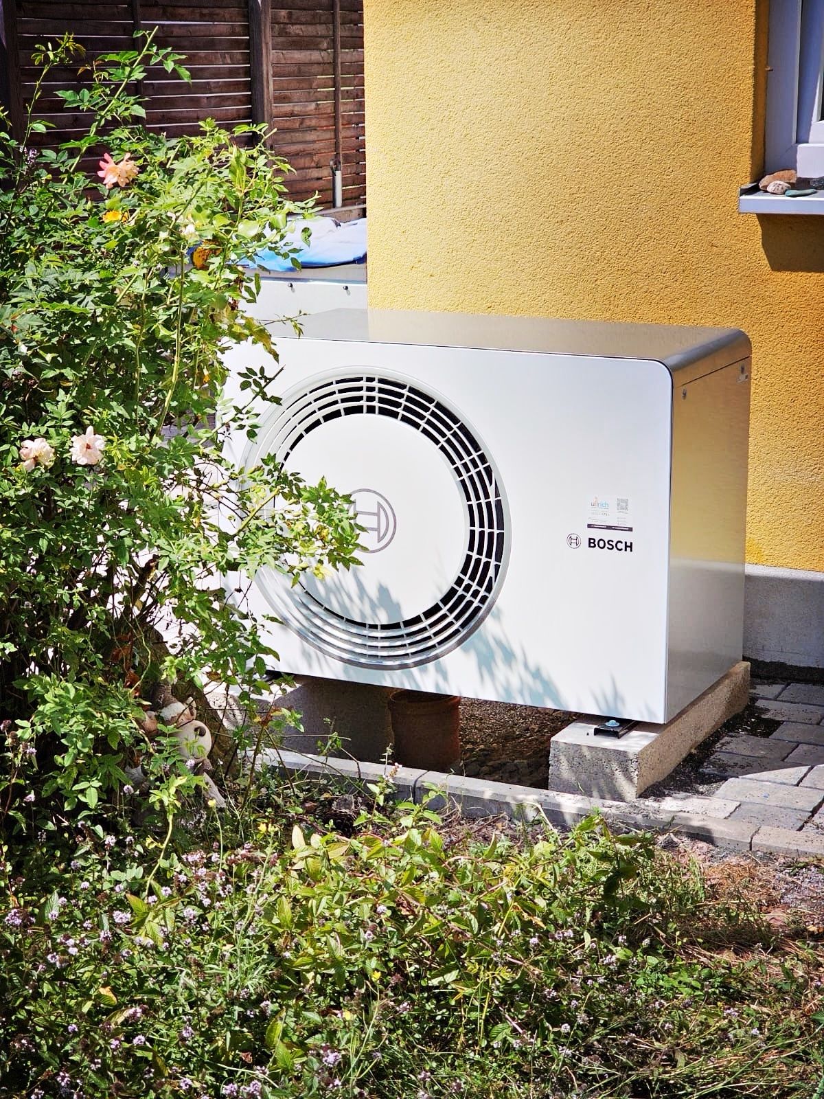 Außeneinheit einer Bosch-Wärmepumpe vor einer gelben Hauswand hinter einem Rosenstrauch