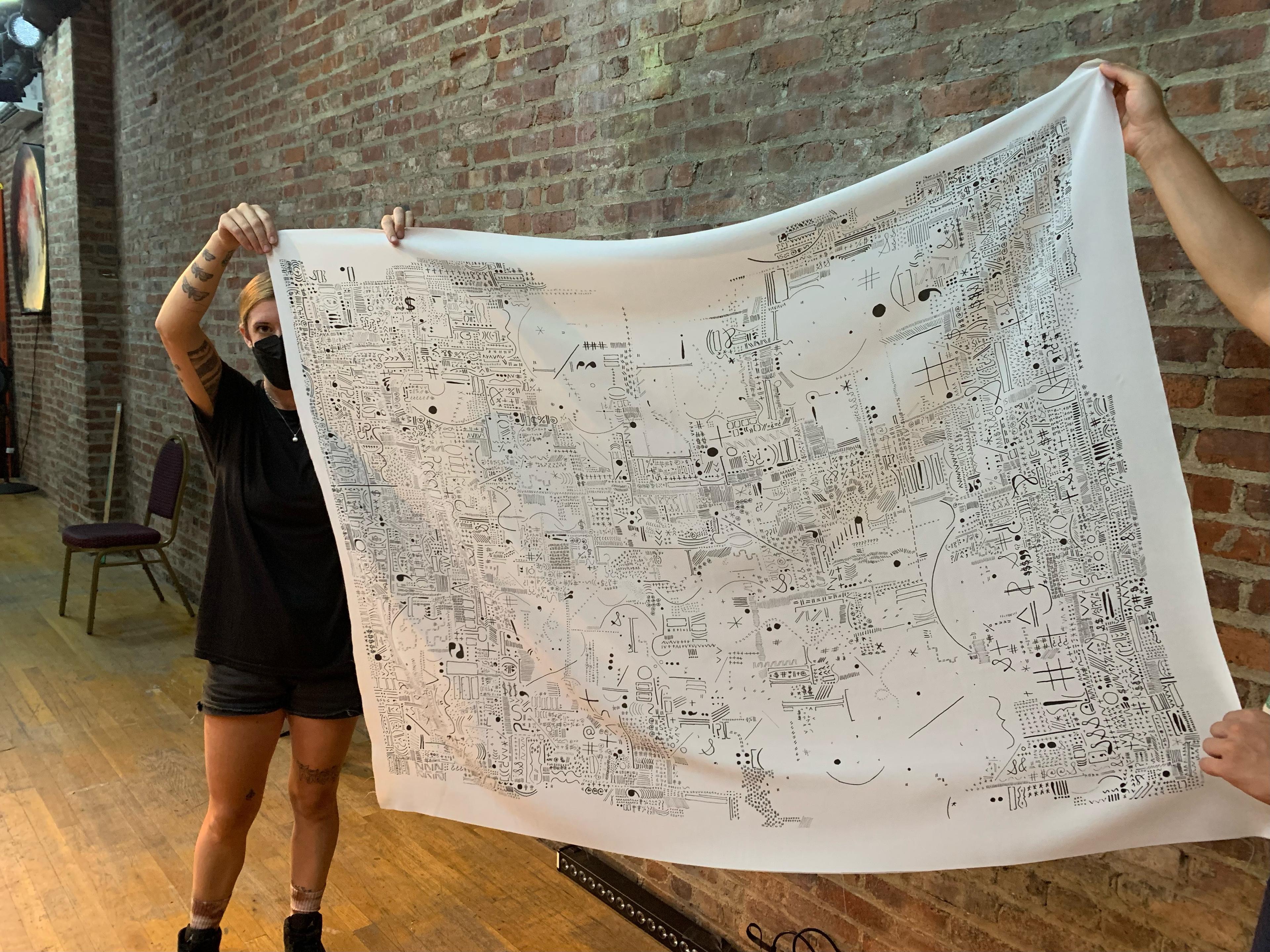 A photograph of Ari, a white person standing and carefully holding the edge a white sheet with small, hand-drawn punctation marks printed on it. Ari wears a black mask with with one looking at the camera and one covered by the tapestry. She has gold hair pulled back and wears short black jean cut-off shorts and a black t-shirt. A brick wall and a chair on a light wood floor is behind her.