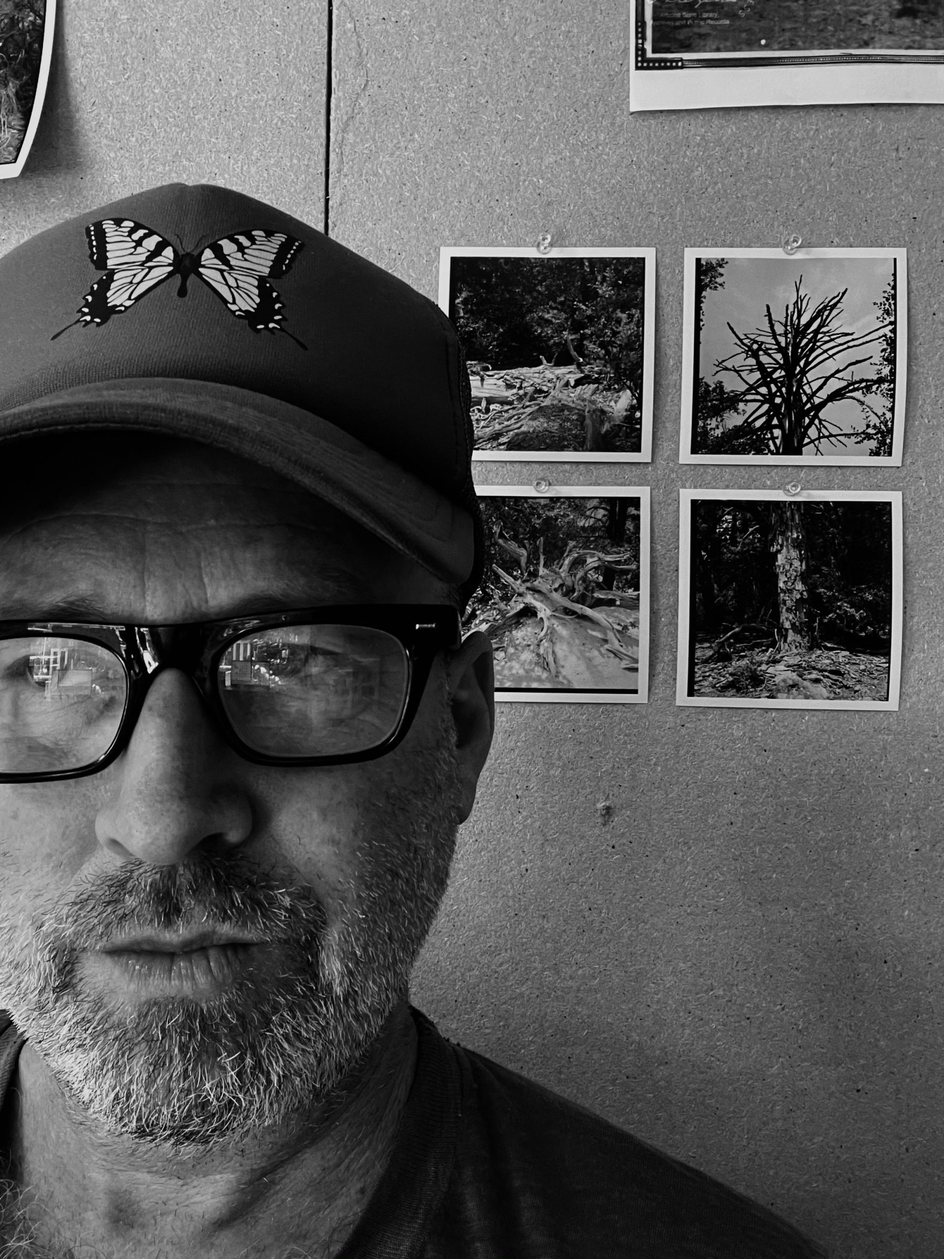 A black and white selfie photo of Ashley Hunt, a white person, pictured in front of a cork board with four photos of trees tacked onto it. He is wearing a hat with a butterfly on it and has a salt and pepper beard.