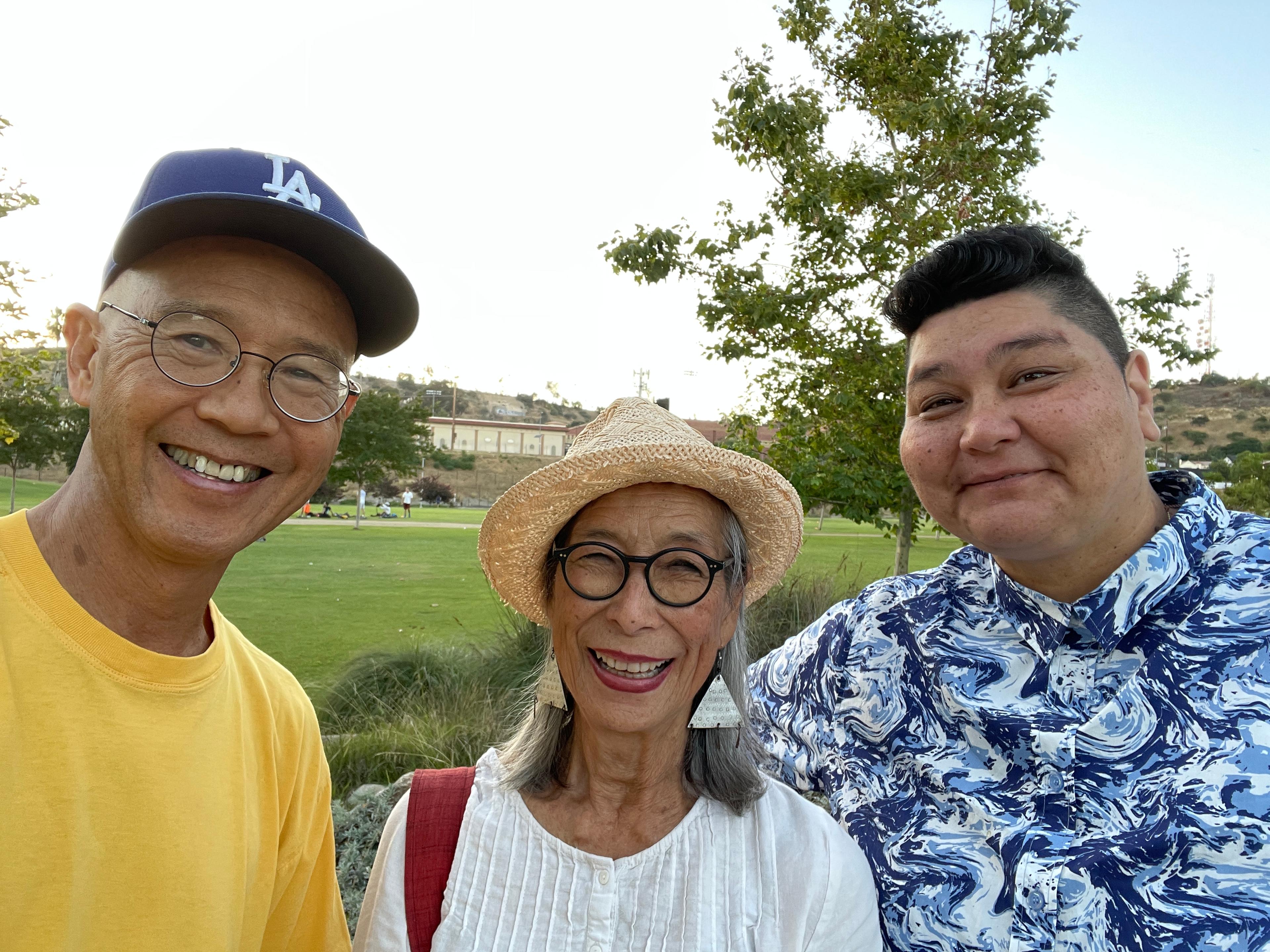 An Asian American man in a yellow shirt and LA Dodgers hat stands next to a smiling elder Japanese American woman in a straw hat, glasses, and white shirt, and a smiling Filipin@e American mixed race gender non-conforming person with a faded haircut and a blue and white shirt.