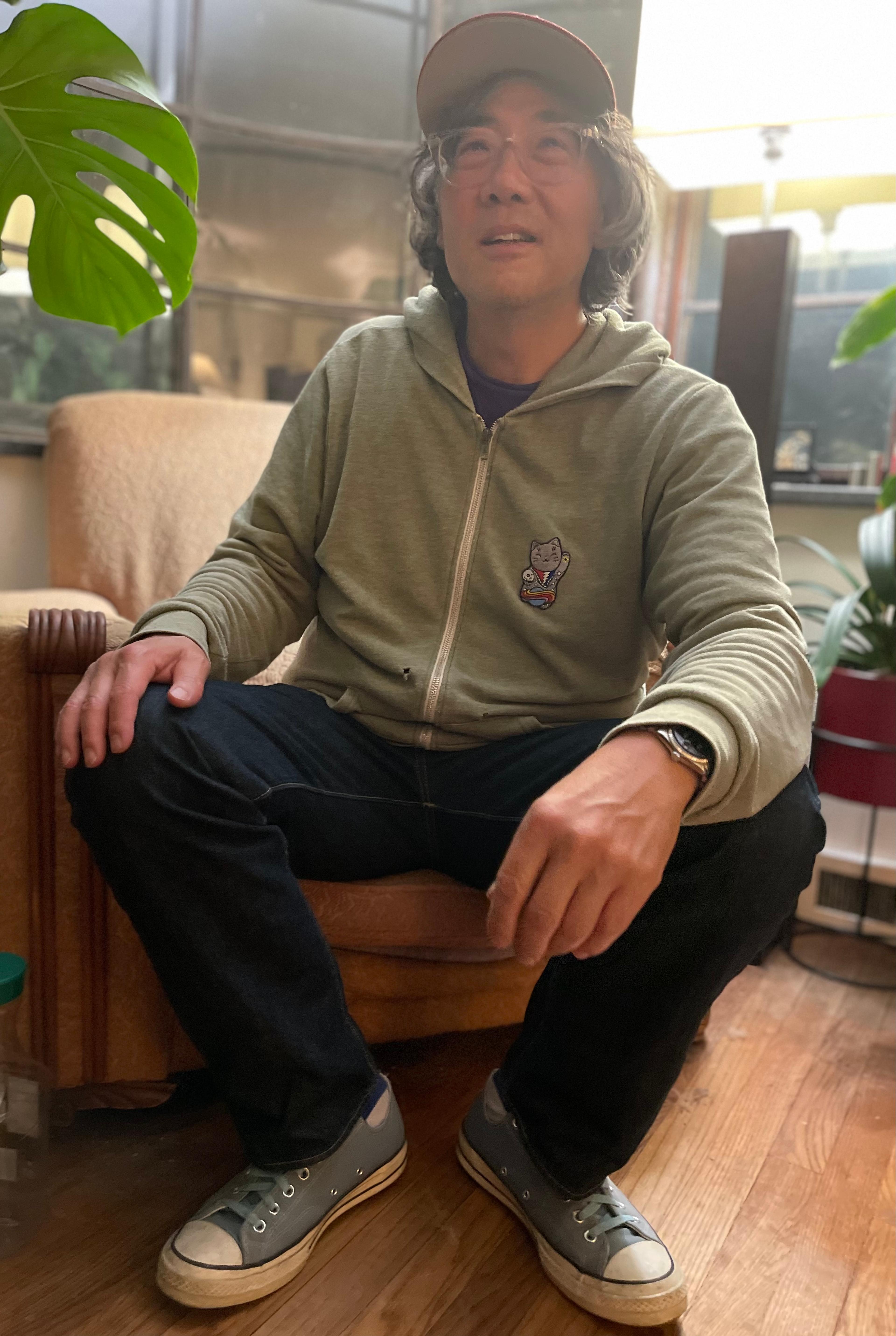 A man wearing a hoodie, Converse sneakers, and baseball cap sits on a chair in a living room. He is photographed from slightly below and there are plants peeking into the frame.