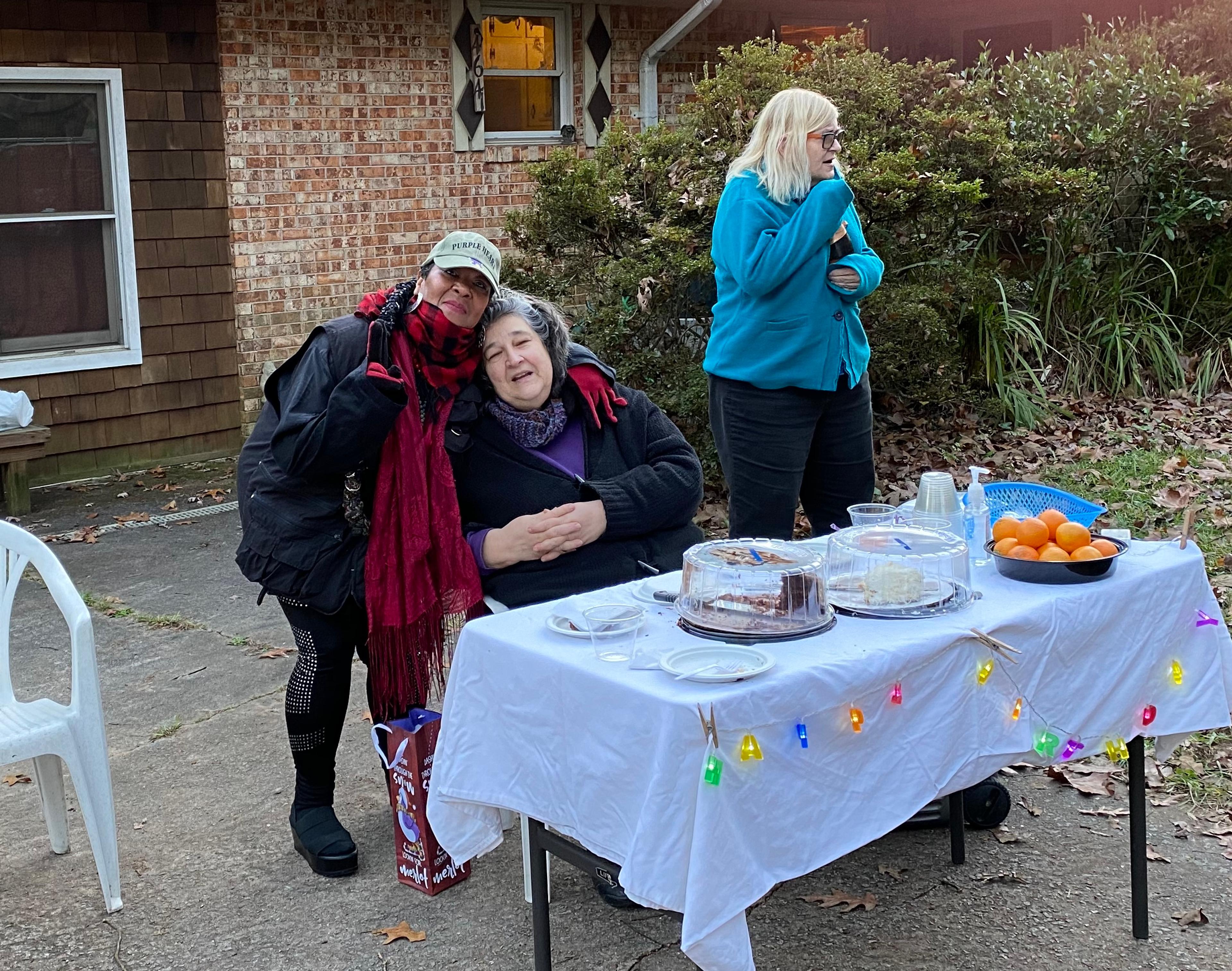 A photo of three people in the driveway of a brick house. They are gathered around a table covered with a white tablecloth decorated with colorful lights. On the table, there are two cakes, a bowl of oranges, and some disposable cups and plates. Kathie, a white person, is sitting at the table. She has gray hair and is wearing a purple shirt and black jacket. A friend stand behind her, smiling at the camera as they embrace Kathie. This friend is wearing a baseball cap, a black jacket, red and black scarf, and black pants with silver studs. On the other side of Kathie, another friend is opening a bottle of champagne. They have blonde hair and are wearing glasses, a blue jacket, and black pants.
