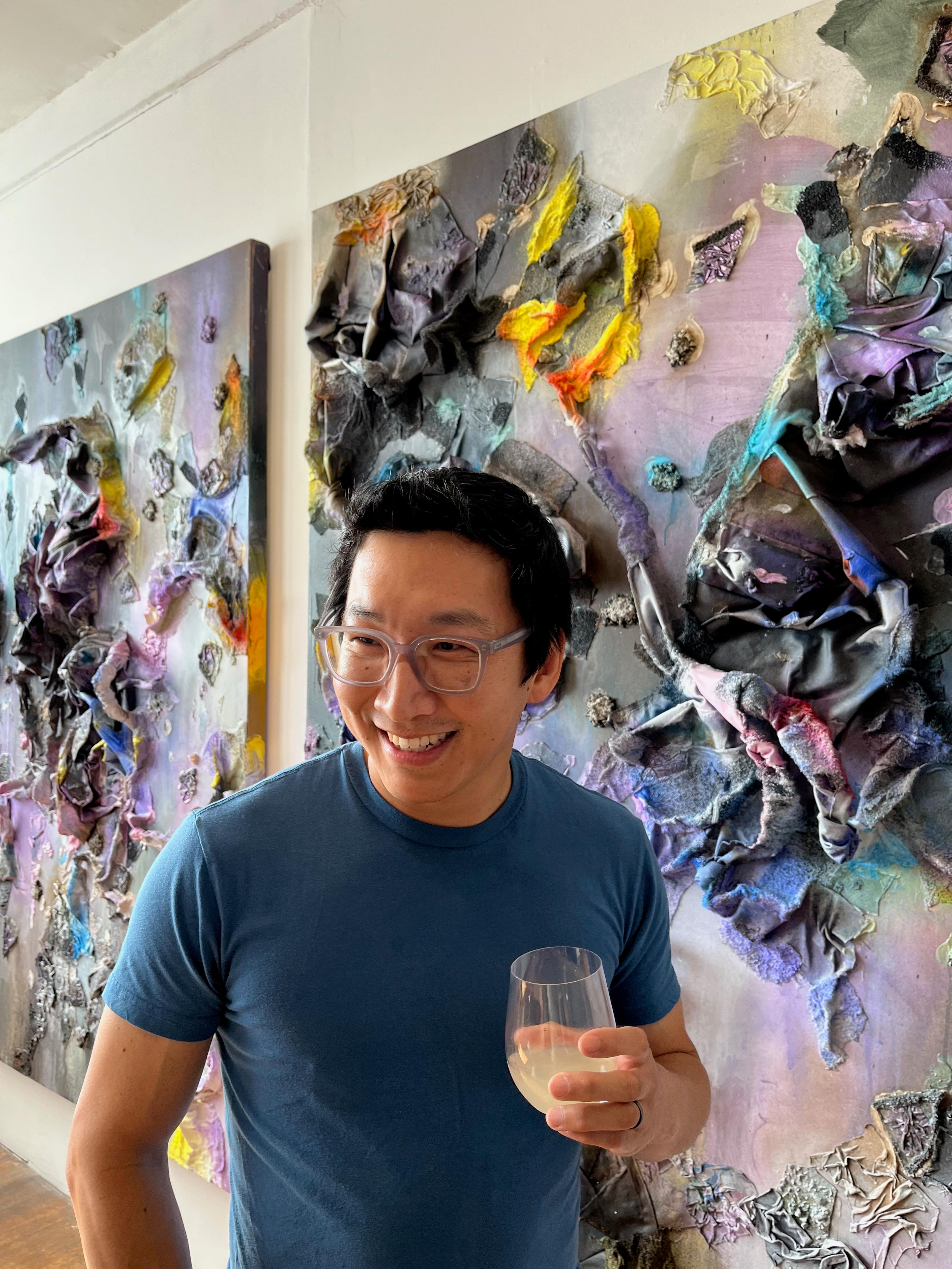 Alex, a Korean man standing and holding a drink while smiling. Alex has glasses and short black hair and wears a blue t shirt. The background includes two paintings.	