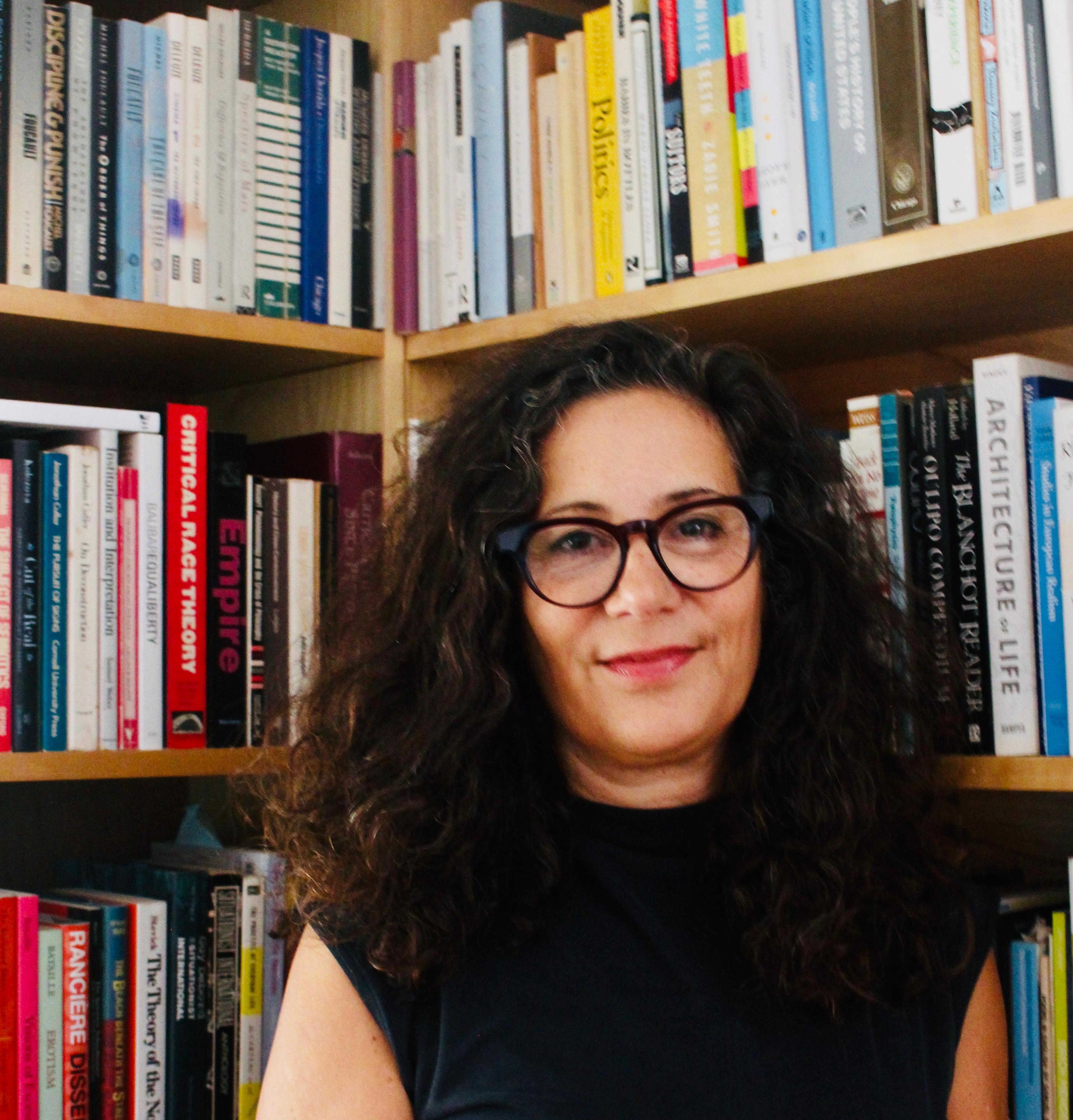 A woman with dark, curly hair and dark-framed glasses stands in front of a bookshelf.