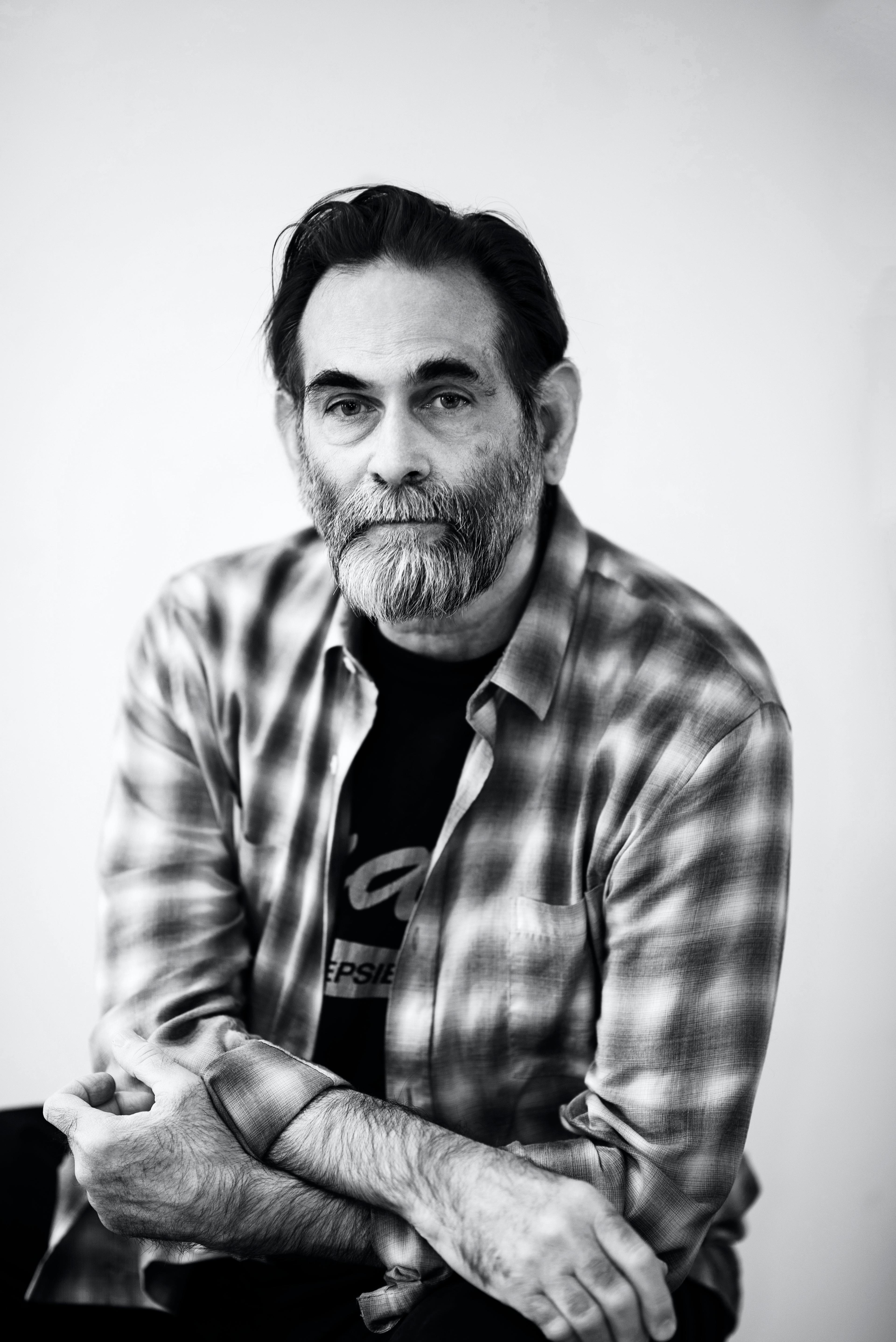 A black and white photo of Avram, a white person, sitting down looking straight at the camera. He has black hair, a salt and pepper beard and is wearing a flannel shirt over a black t-shirt.